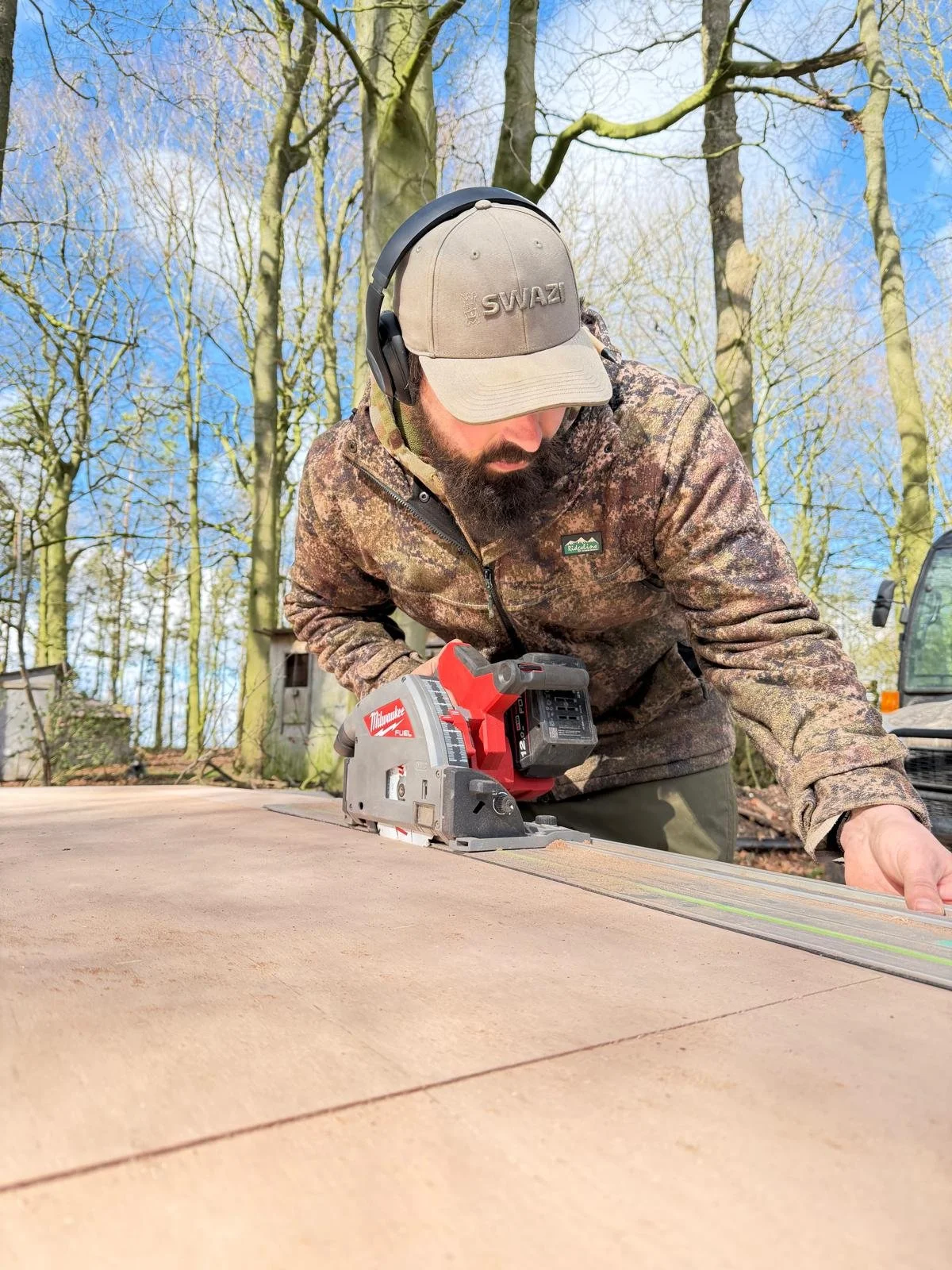 A man in camouflage jacket, wearing a tan cap with "SWAZI" embroidered on it, working outdoors with a power saw on a wooden surface, surrounded by trees and construction equipment.