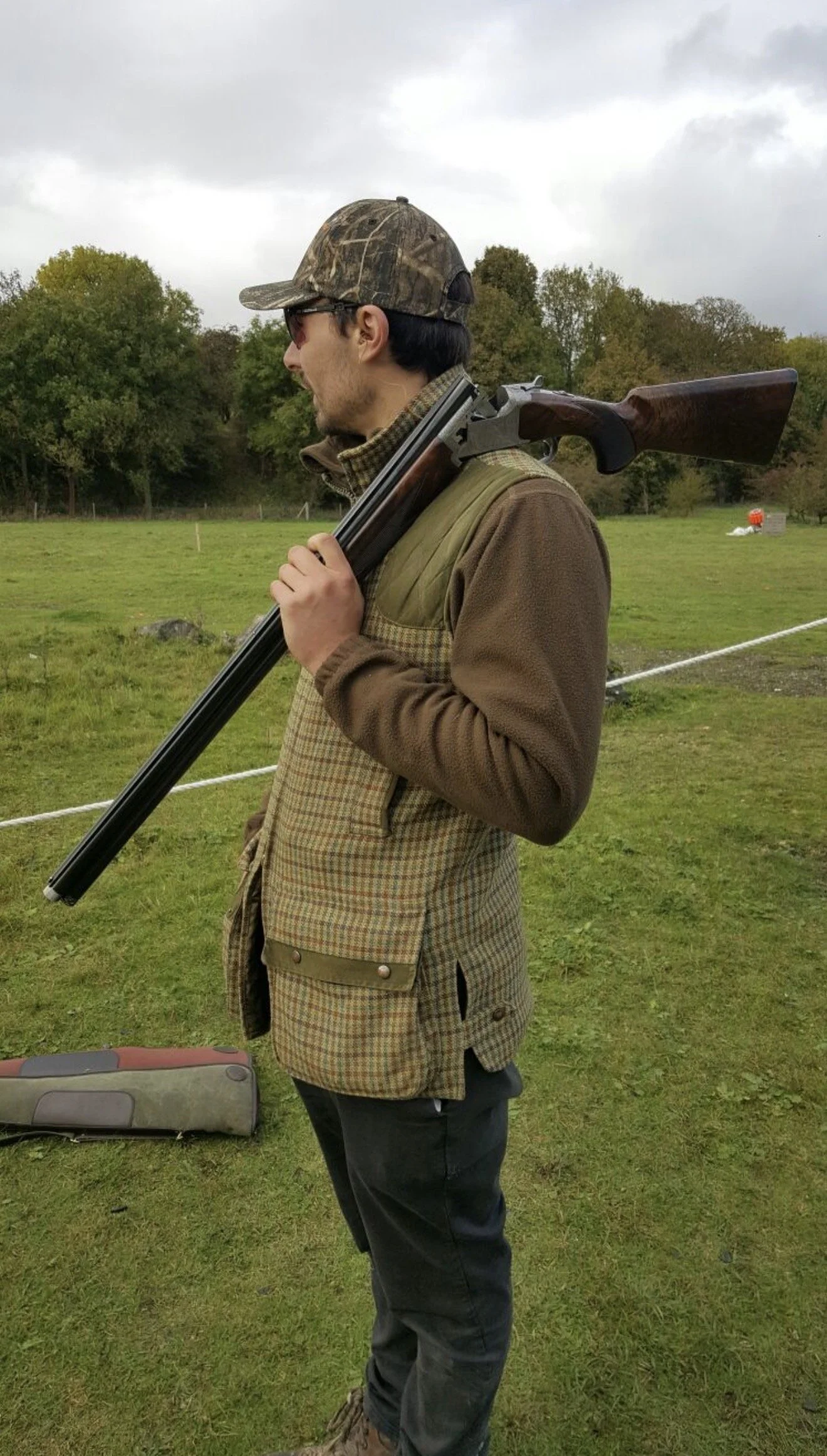 A man in camouflage cap, sunglasses, and a brown jacket with a checked vest over it, standing outdoors on grass, carrying a shotgun over his shoulder.