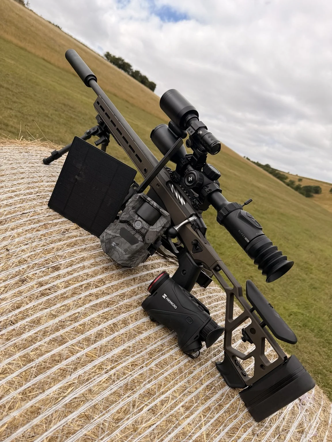 A tactical rifle with a scope and accessories resting on a hay bale in an open field.