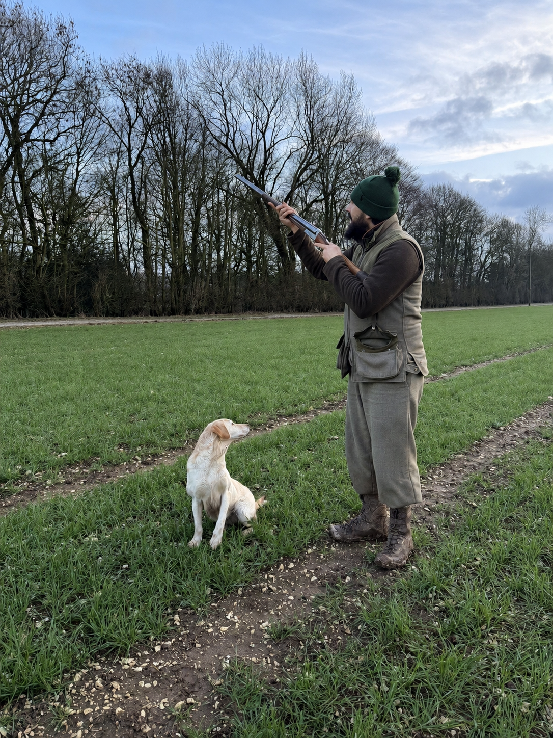 A man in outdoor hunting gear, including a green beanie and boots, aiming a shotgun at the sky while a yellow and white dog sits nearby on a grassy field with trees in the background.