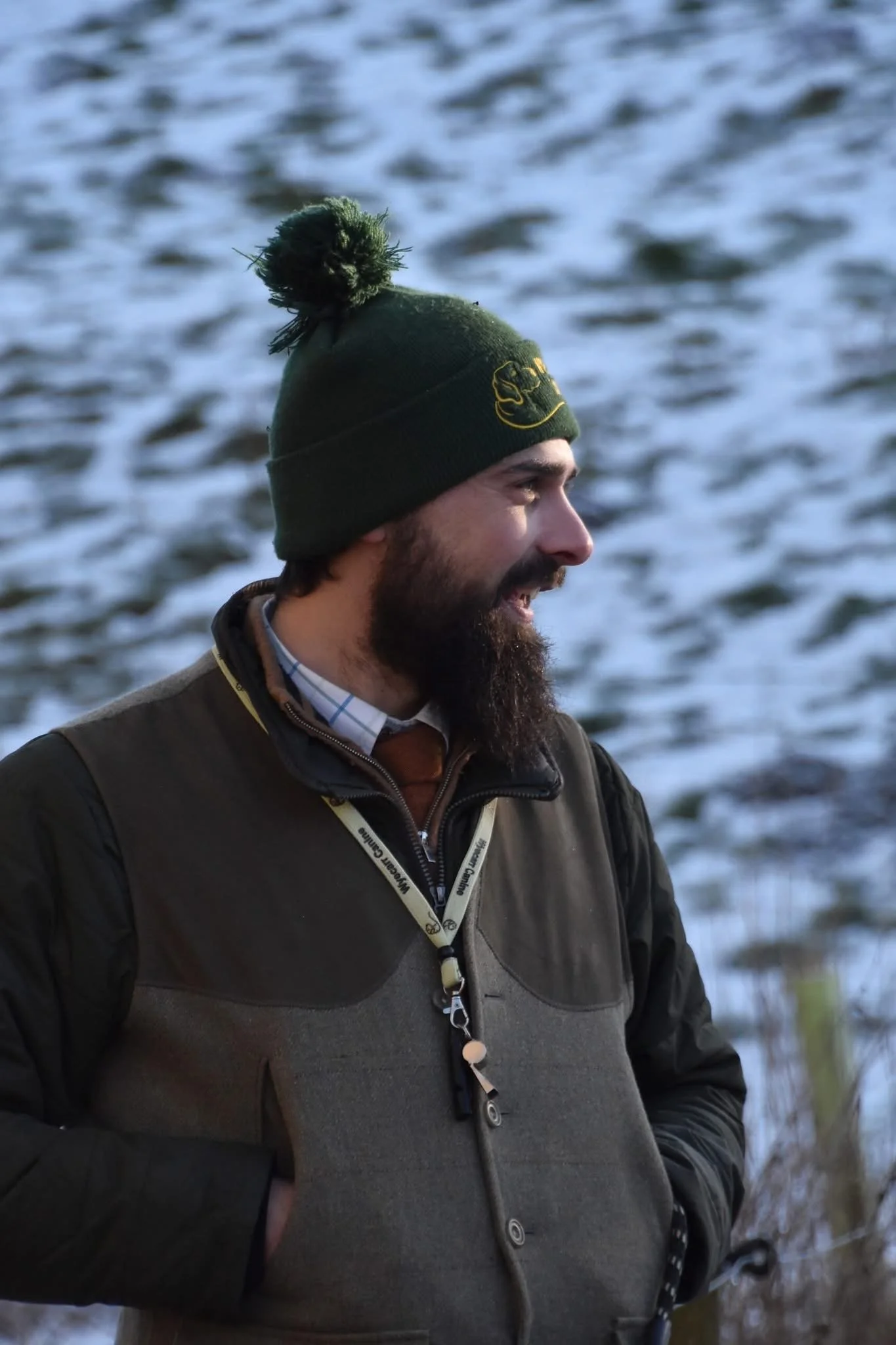 A man with a beard is standing outdoors near a body of water, wearing a green knit hat with a pom-pom and a dark jacket.