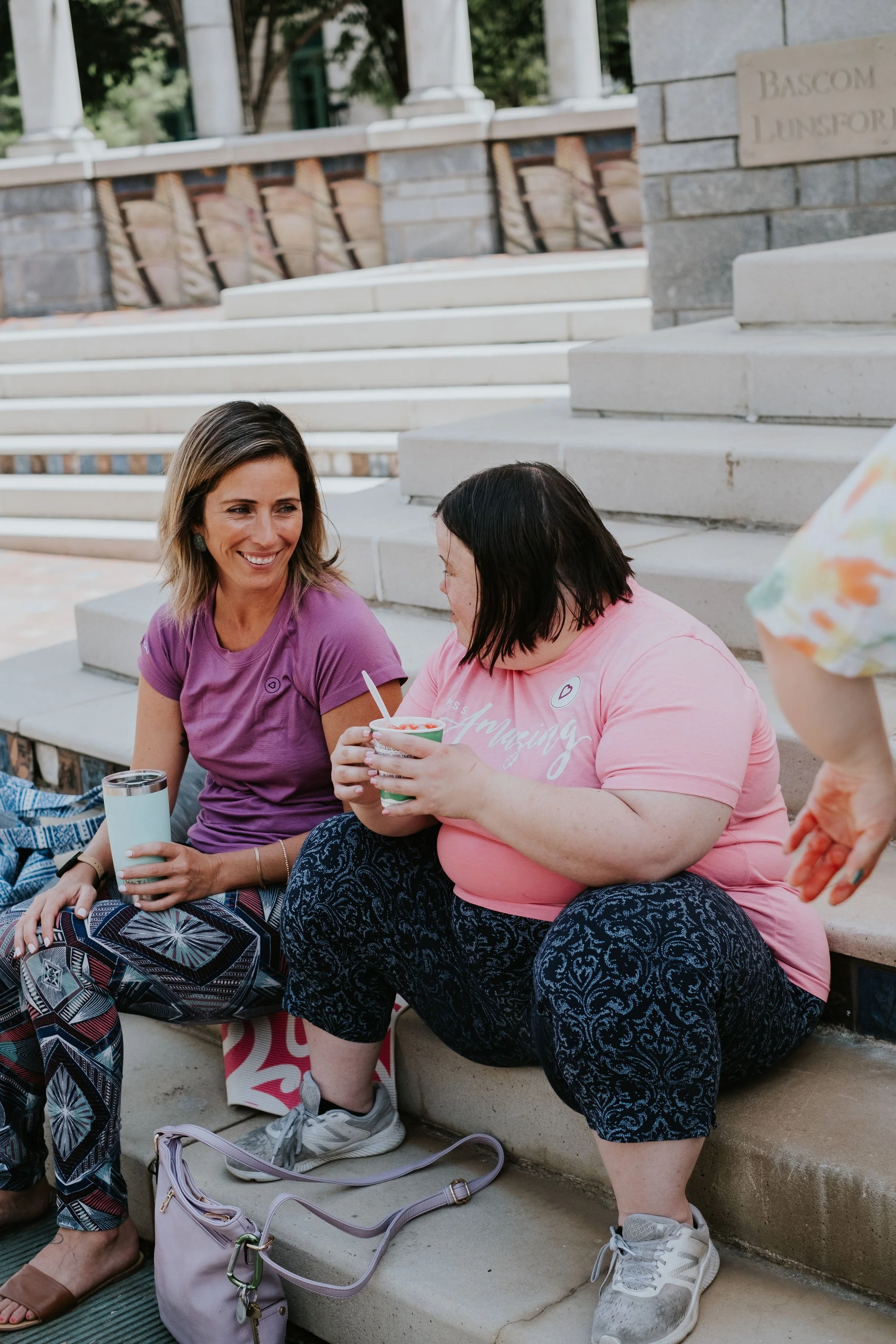 Two women sitting on steps outdoors, smiling and talking, one holding a cup of yogurt, the other holding a spoon, with backpacks and personal items on the ground beside them.