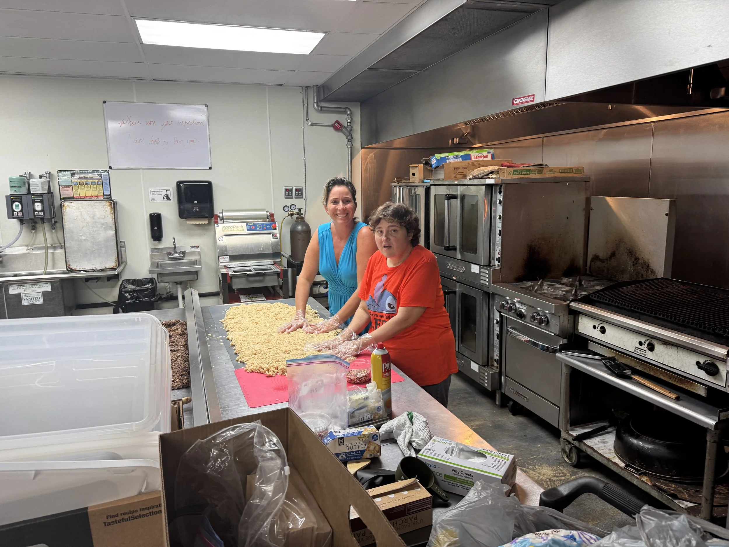 Two women are working together in a commercial kitchen, preparing food on a stainless steel counter. They are smiling and wearing gloves. The kitchen has various appliances and supplies, including an oven, a large industrial range, and boxes of food ingredients.