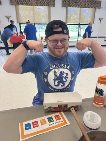 A smiling man wearing glasses, a black cap backwards, and a Chelsea Football Club t-shirt flexing his arms at a table in a bright room with large windows and other people in the background.