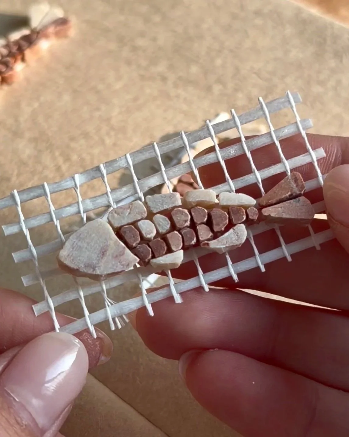 Close-up of a miniature, handmade mosaic on a small piece of mesh, with various small stones and tiles attached, held between hands.