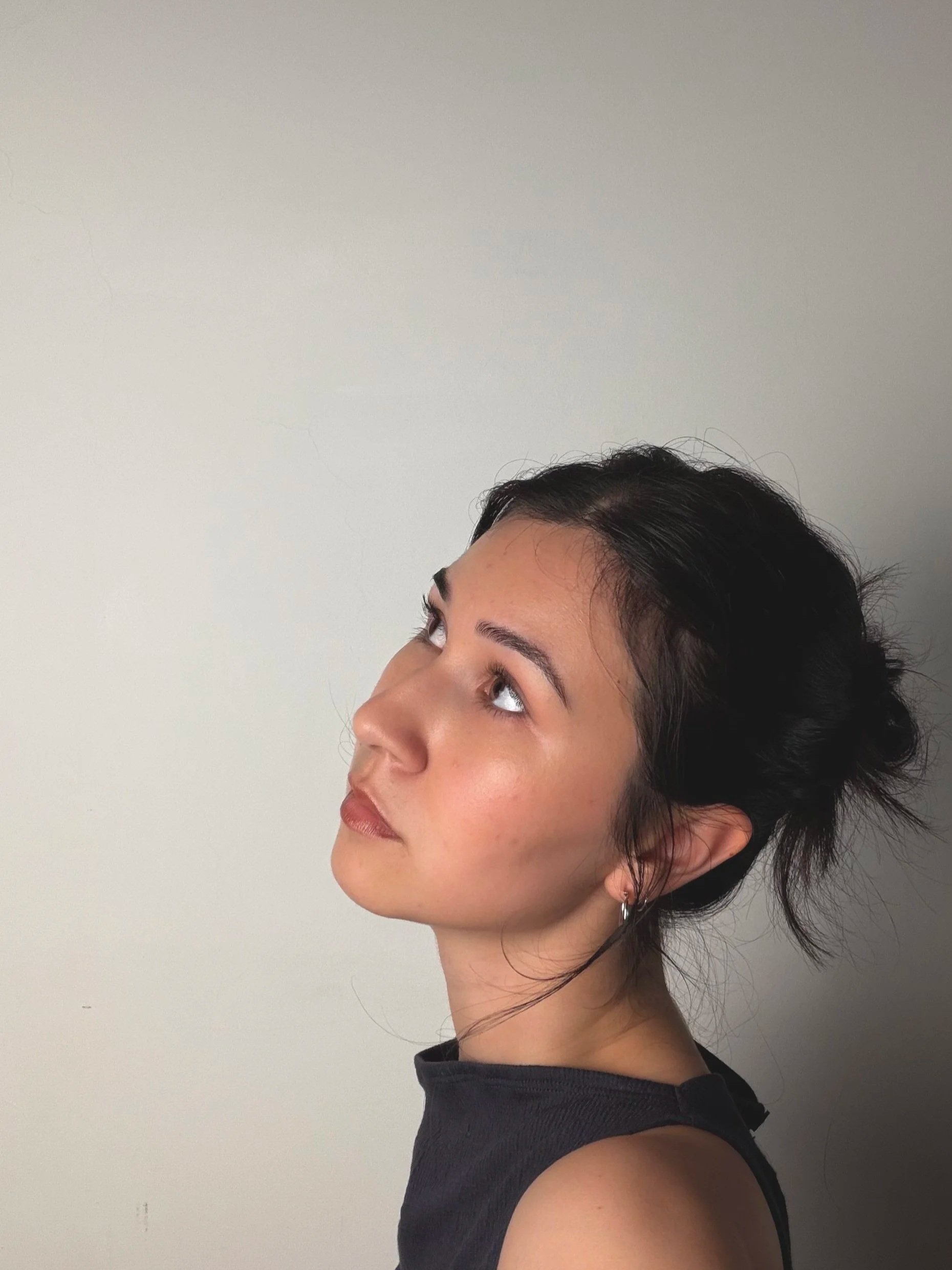 A woman with dark hair styled in a messy fashion, wearing a black sleeveless top and earrings, looking upward against a plain light-colored background.