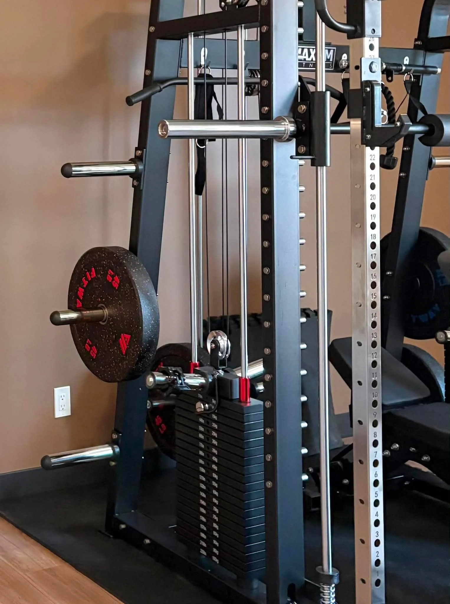 A home gym setup with a rack holding weights, a weight plate on the ground, a weight stack, and various exercise attachments against a brown wall with a power outlet.
