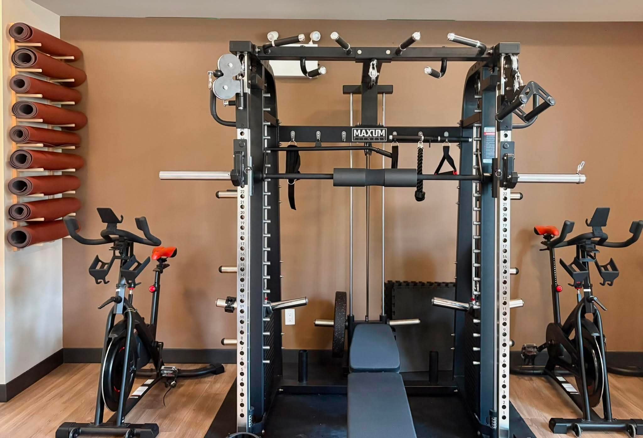 A gym with various fitness equipment including a cable machine, stationary bikes, a rowing machine, and yoga mats stored on a wall rack.