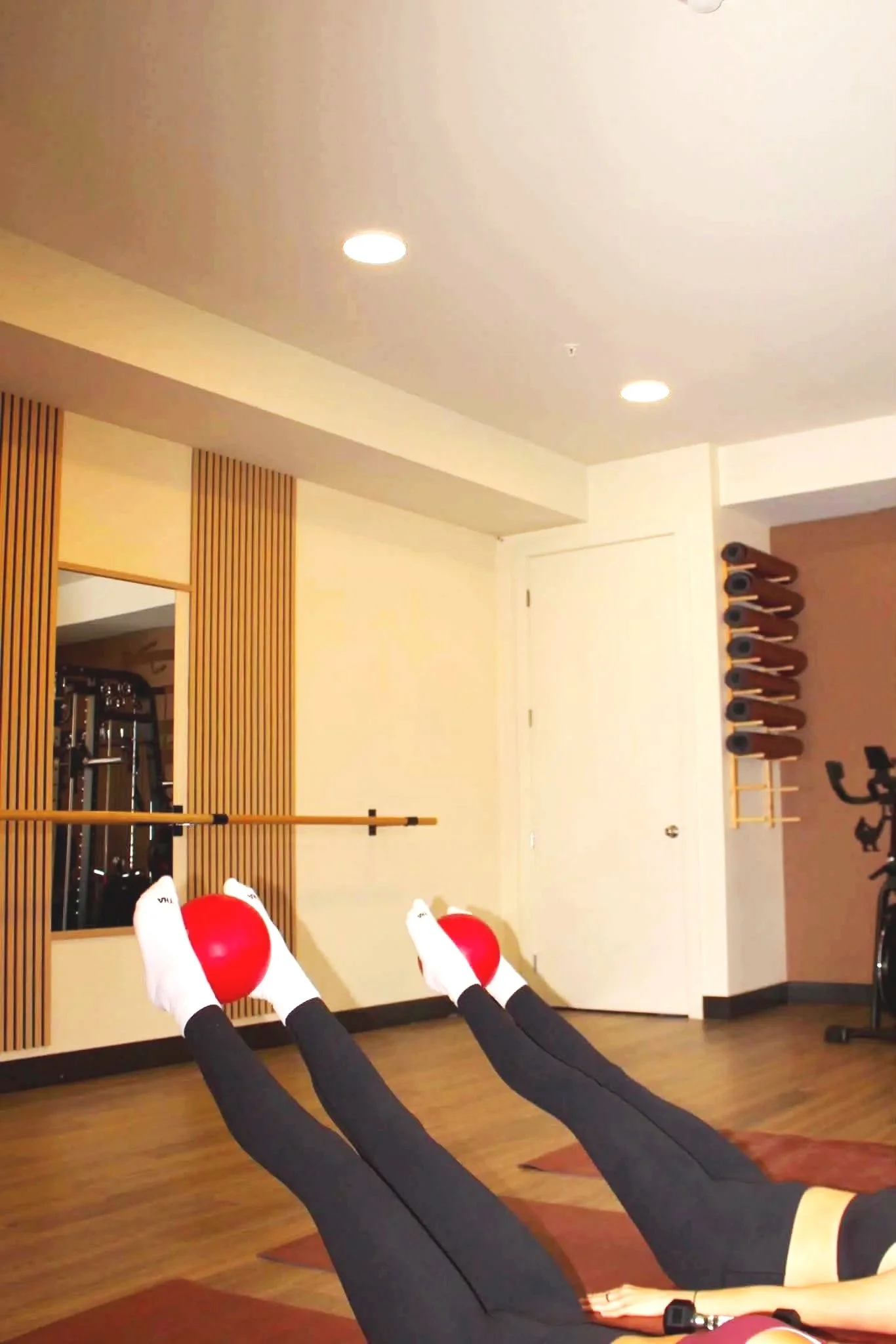 People practicing yoga or stretching with legs extended, feet wearing white socks with black text, on yoga mats in a fitness studio. The room has wooden flooring, a wall with vertical wooden slats, exercise equipment, and teal yoga mats rolled up on a rack.
