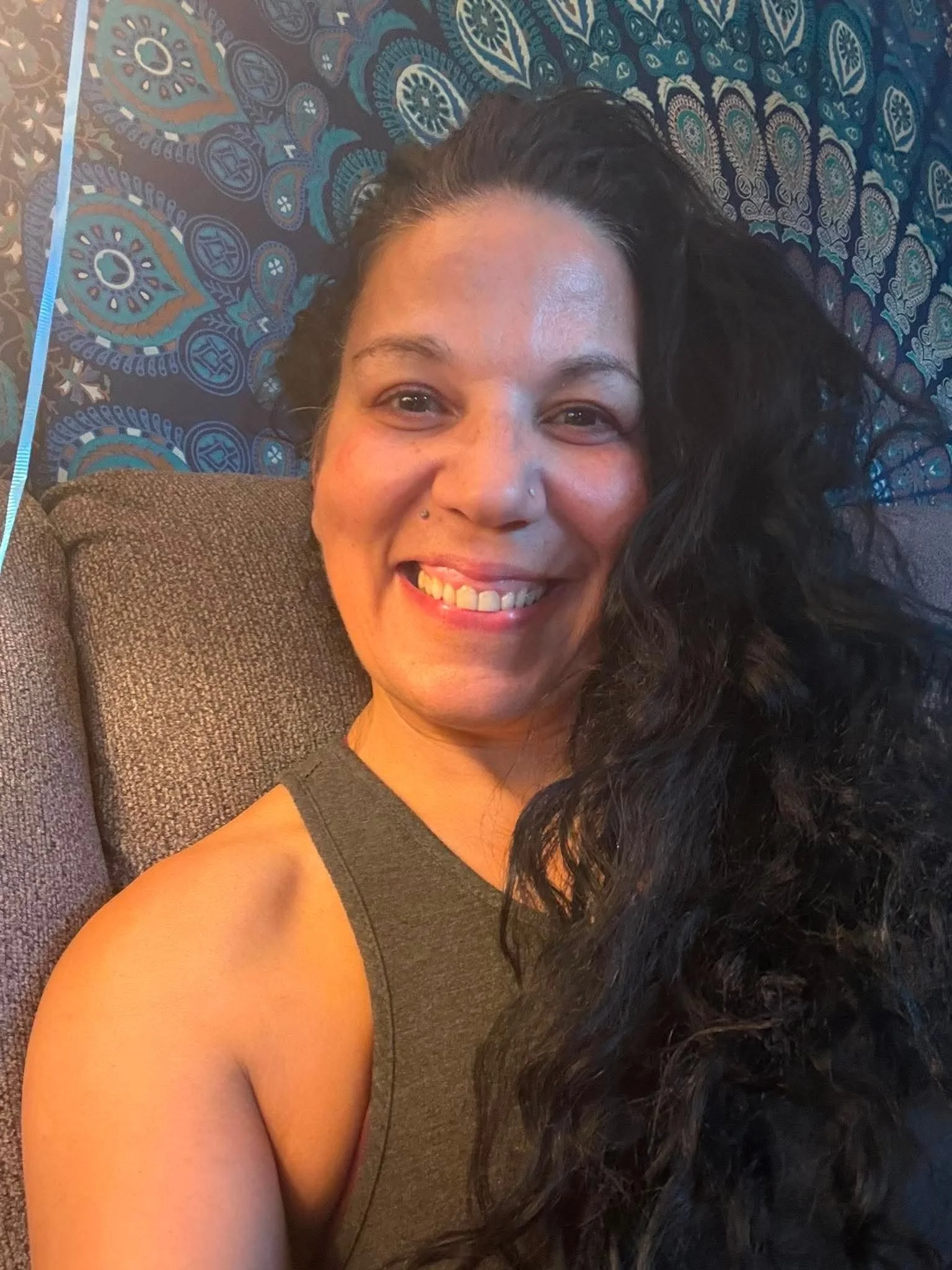 A woman with long dark curly hair, smiling, lying on a gray couch with a patterned blanket behind her.