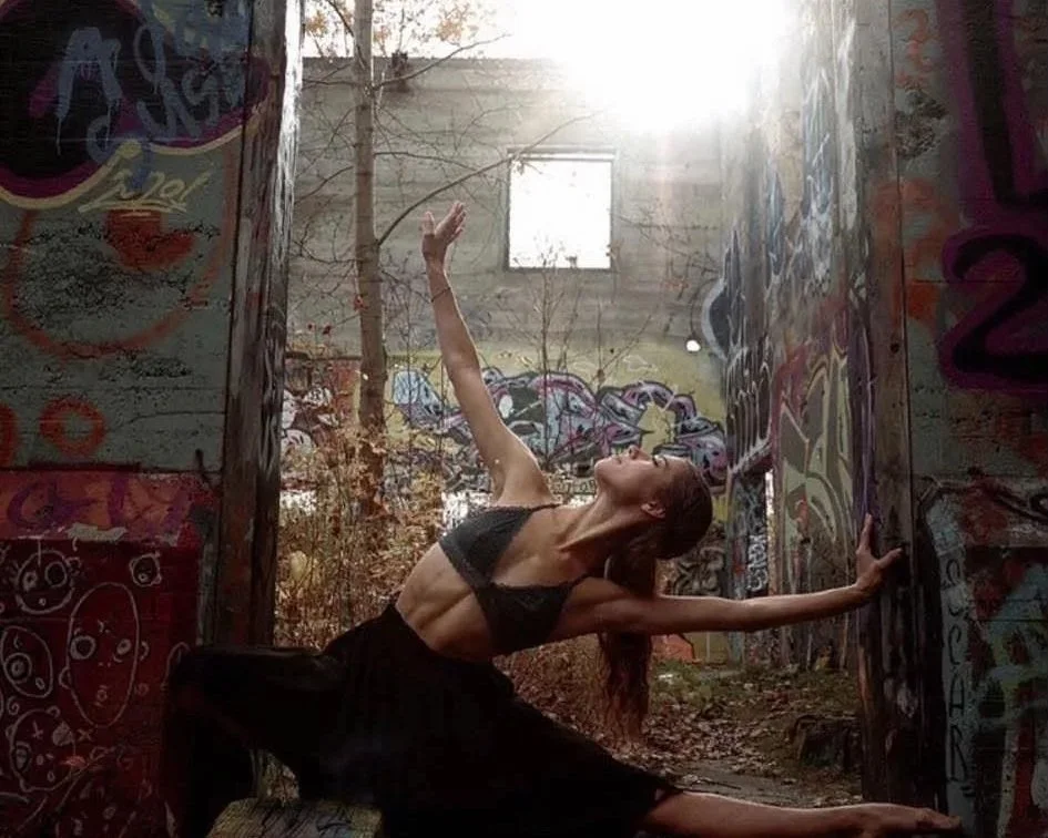 A woman in athletic clothing performs a dance or yoga pose in a graffiti-covered abandoned building with sunlight streaming through an open window.