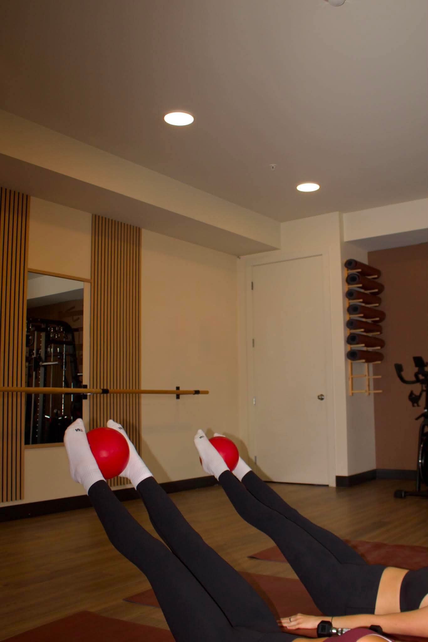Person exercising on a yoga mat with legs elevated, holding a small red exercise ball between their legs, in a studio with wooden flooring and wall paneling.