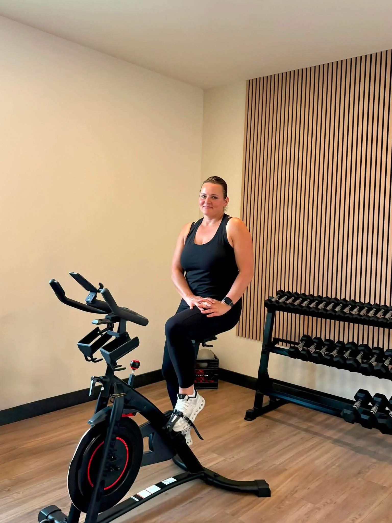 A woman in workout clothes sitting on a stationary exercise bike in a home gym with wooden flooring, a beige wall, and a brown slatted wall panel, next to a multi-dumbbell rack.