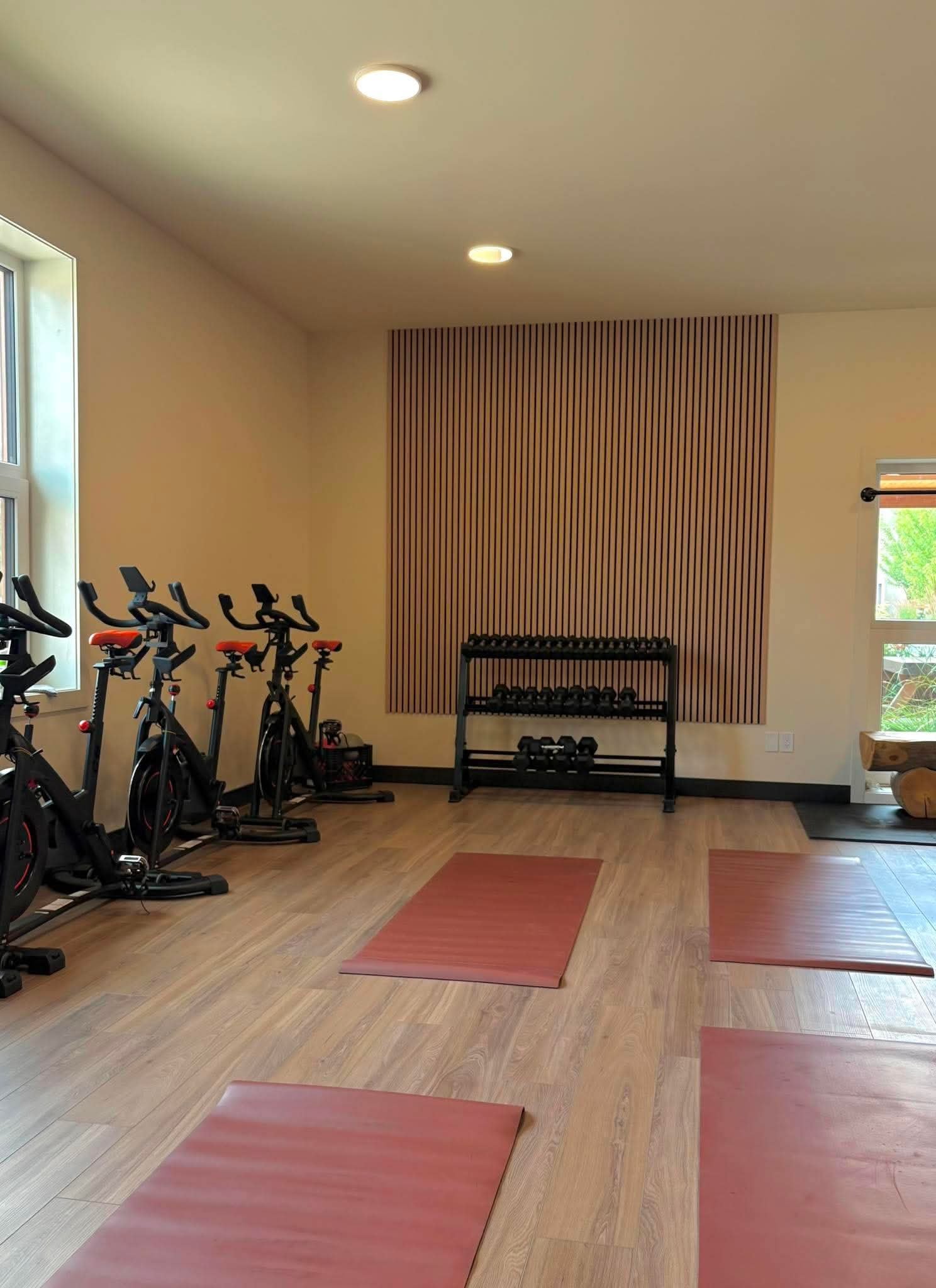 An interior gym with exercise bikes, a weight rack, and yoga mats on wooden floors, illuminated by ceiling lights and a window with natural light.