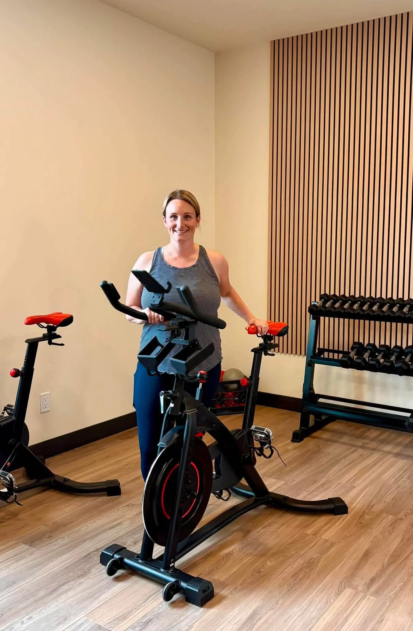 A woman in workout clothing standing next to an exercise bike in a gym or fitness room with beige and brown walls and wood flooring.