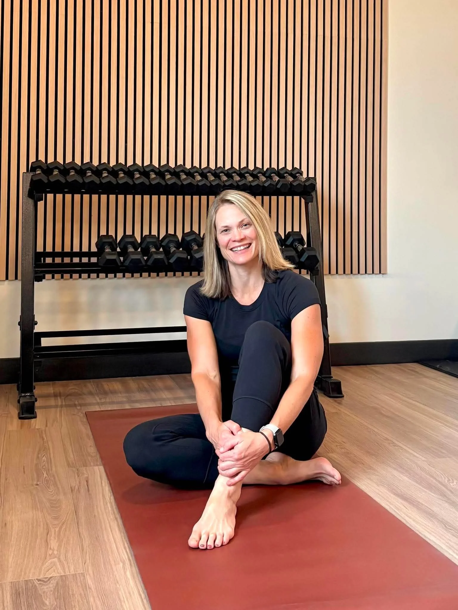 A woman with blonde hair sitting on a yoga mat, smiling, in front of a wooden wall with gym equipment in the background.