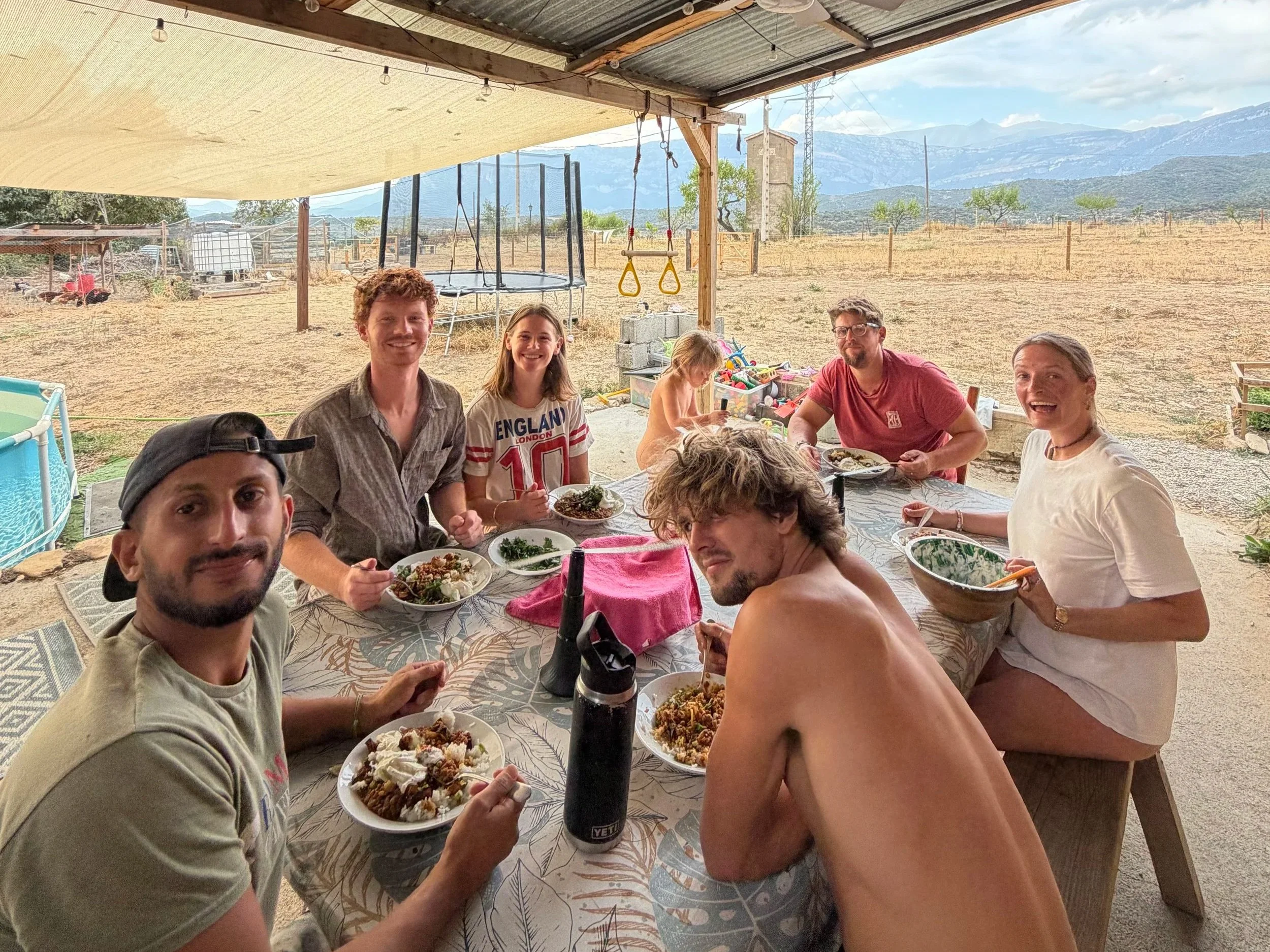Group of seven people sitting and standing around a table outdoors, enjoying a meal together with plates of food and drinks, with a rural landscape and mountains in the background.