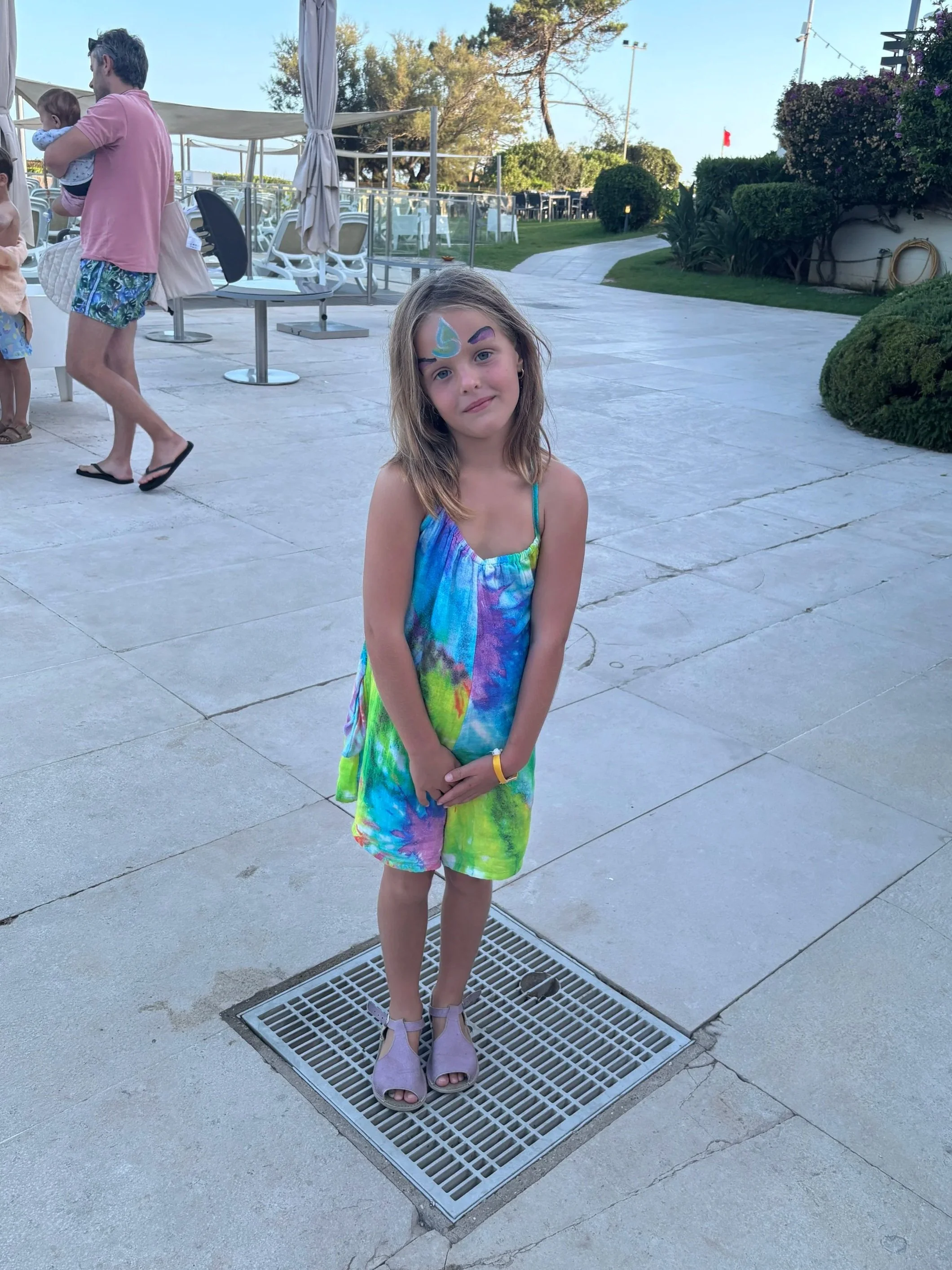 A young girl in a colorful tie-dye dress stands on a drain grate outdoors at a recreational area. She has pastel face paint on her forehead and purple eyebrows, and wears light purple shoes. In the background, there are lounge chairs, tables, green bushes, and a sidewalk, with other people behind her.