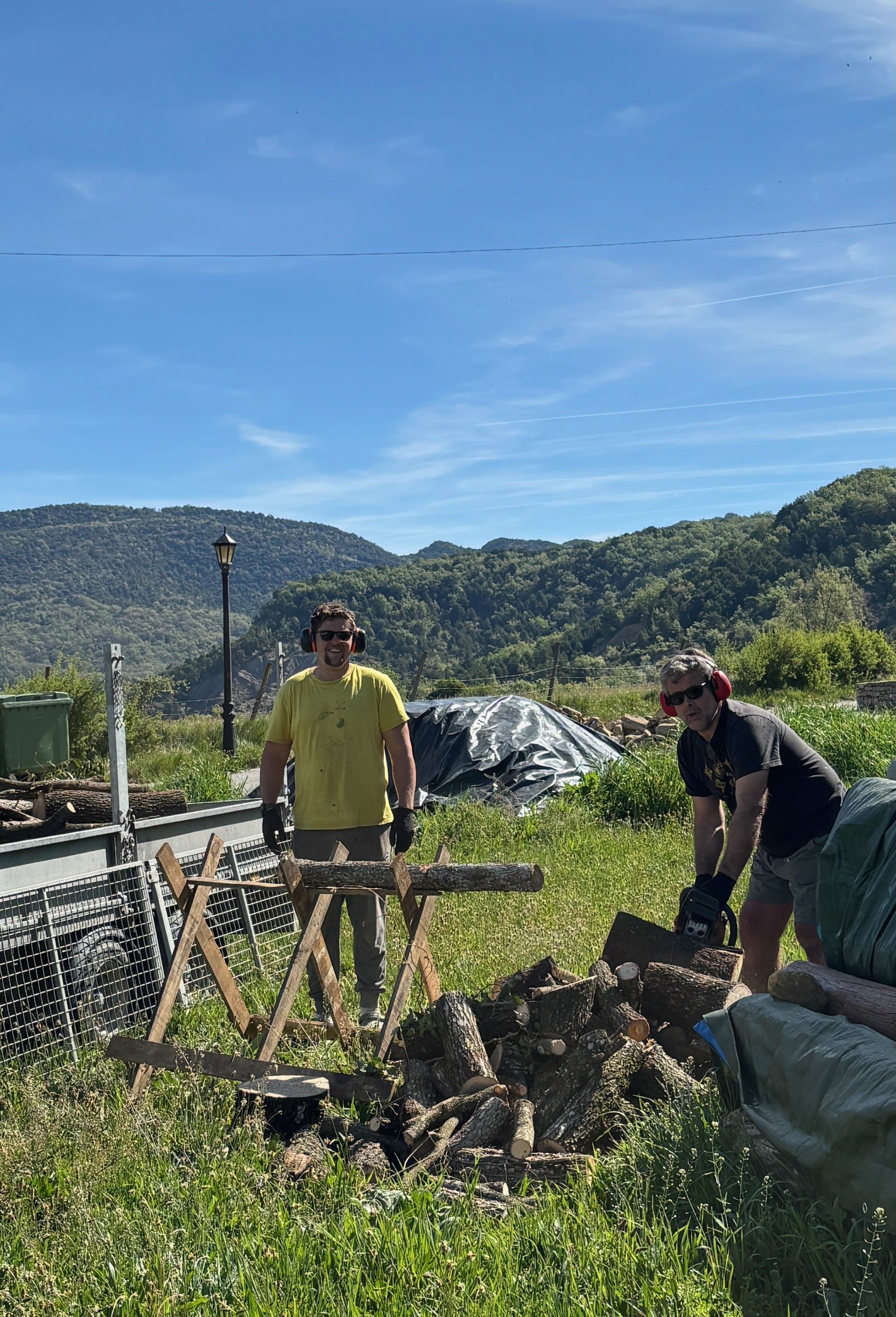 Two men working outdoors with logs, fencing, and tools, in a sunny field with green hills and a blue sky.
