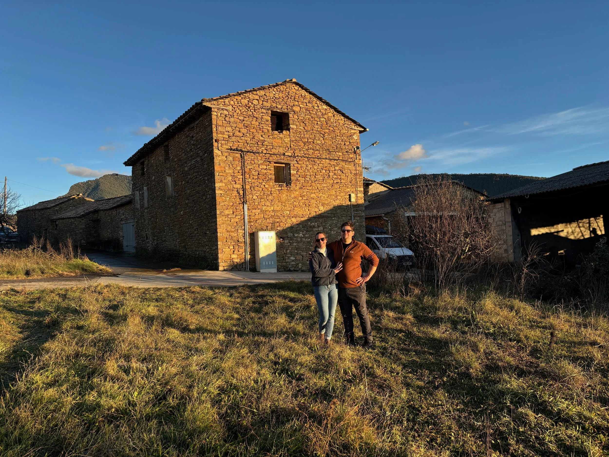 A stone house with a tiled roof during sunset, with a man and woman standing on grass in front, smiling and posing for the camera. Mountains are visible in the background.