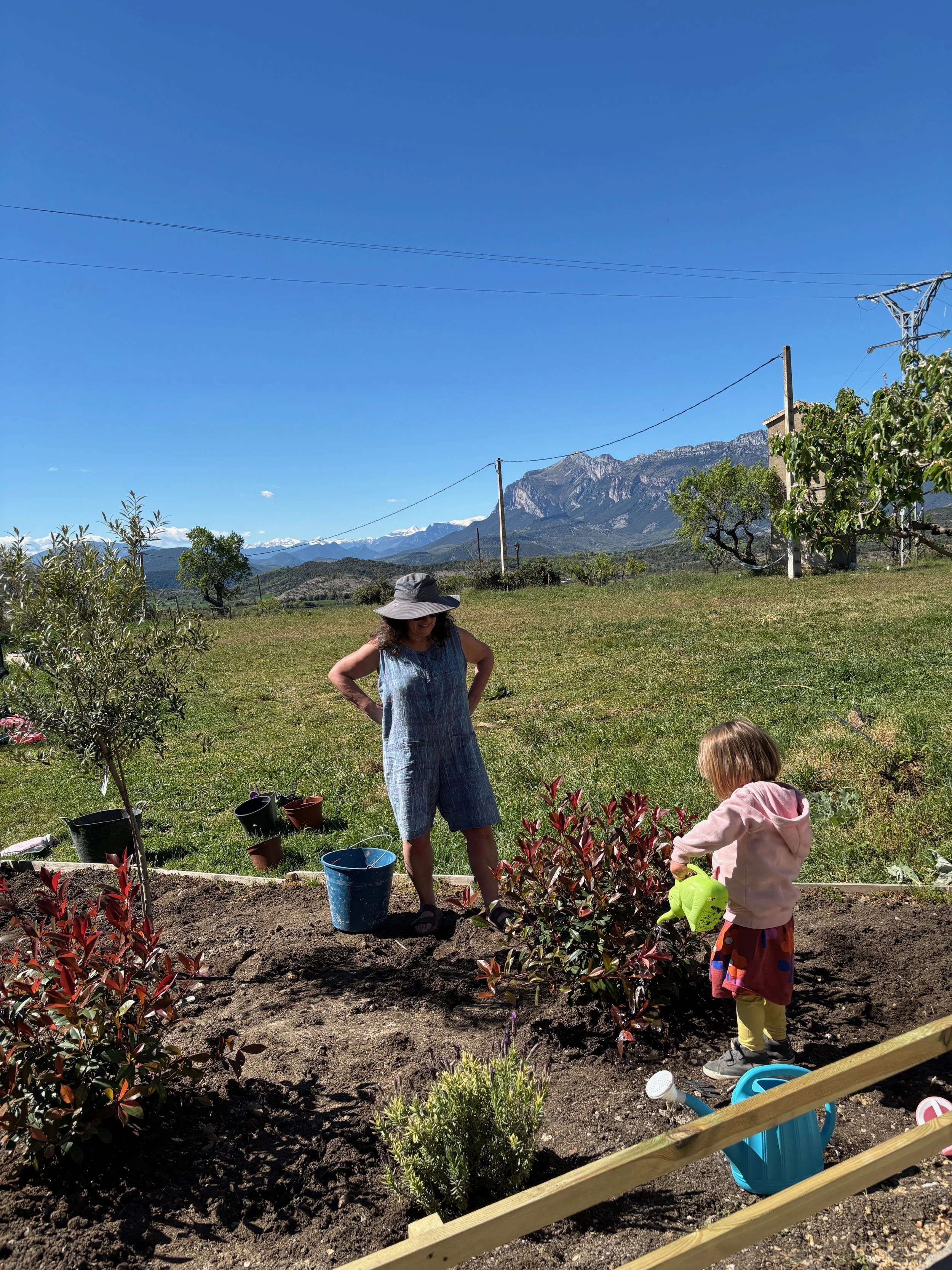 A woman and a young girl planting flowers in a garden under a clear blue sky with mountains in the background.