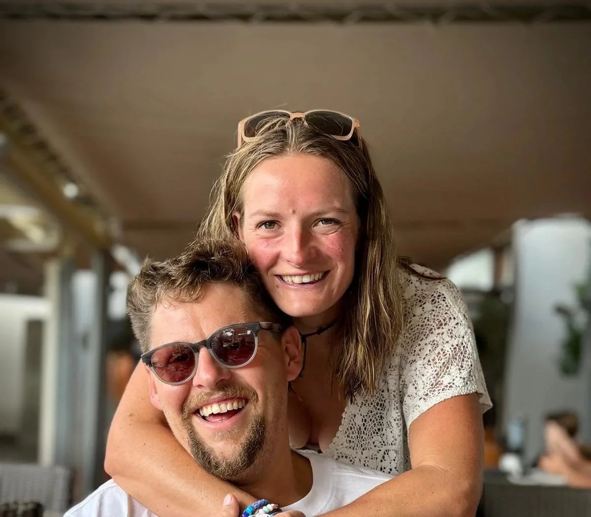 A smiling woman with sunglasses on her head and a man with sunglasses, both enjoying a moment together, with the woman hugging the man from behind.