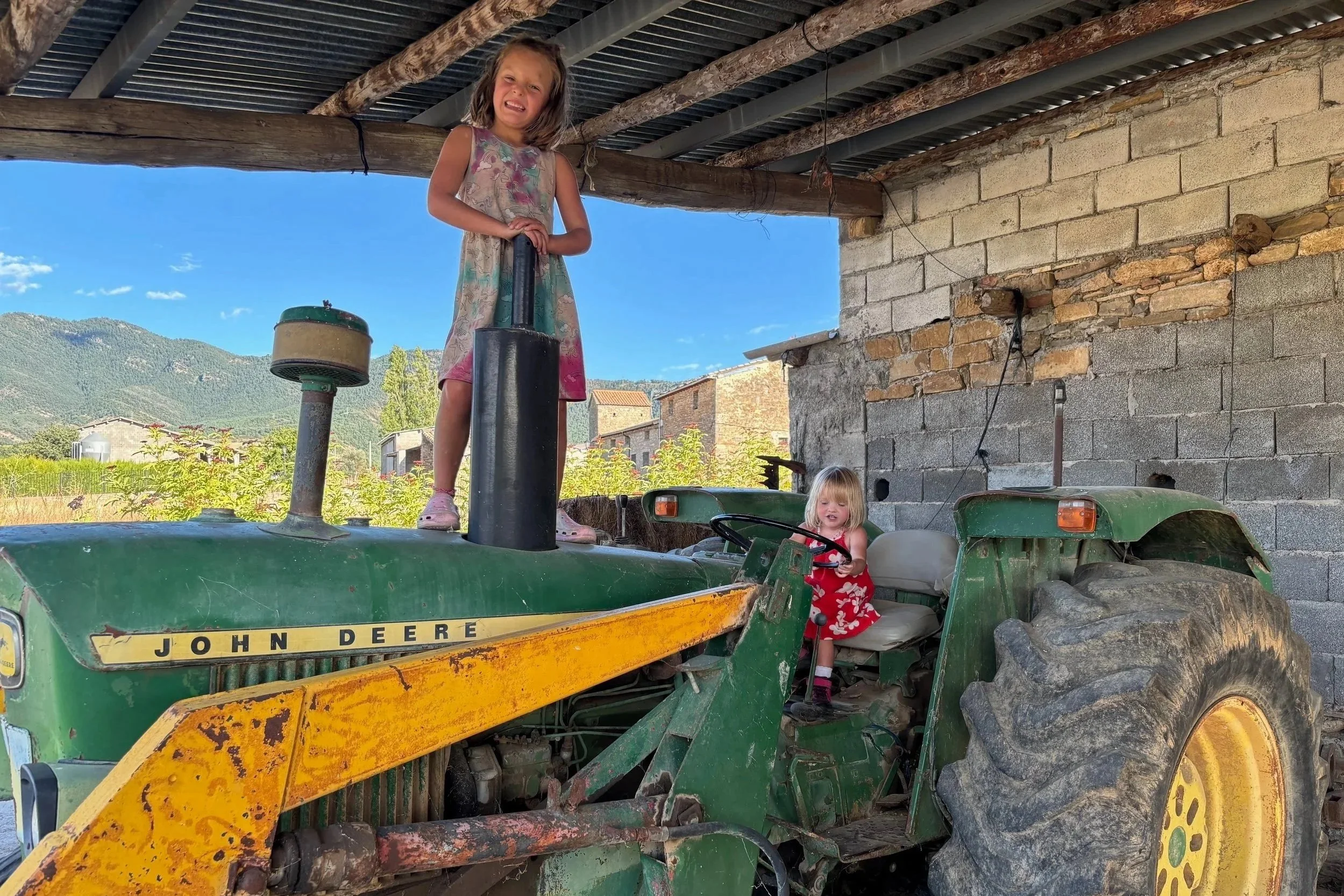 Two young girls playing on a green John Deere tractor inside a barn or shed, with a mountain landscape visible outside on a sunny day.