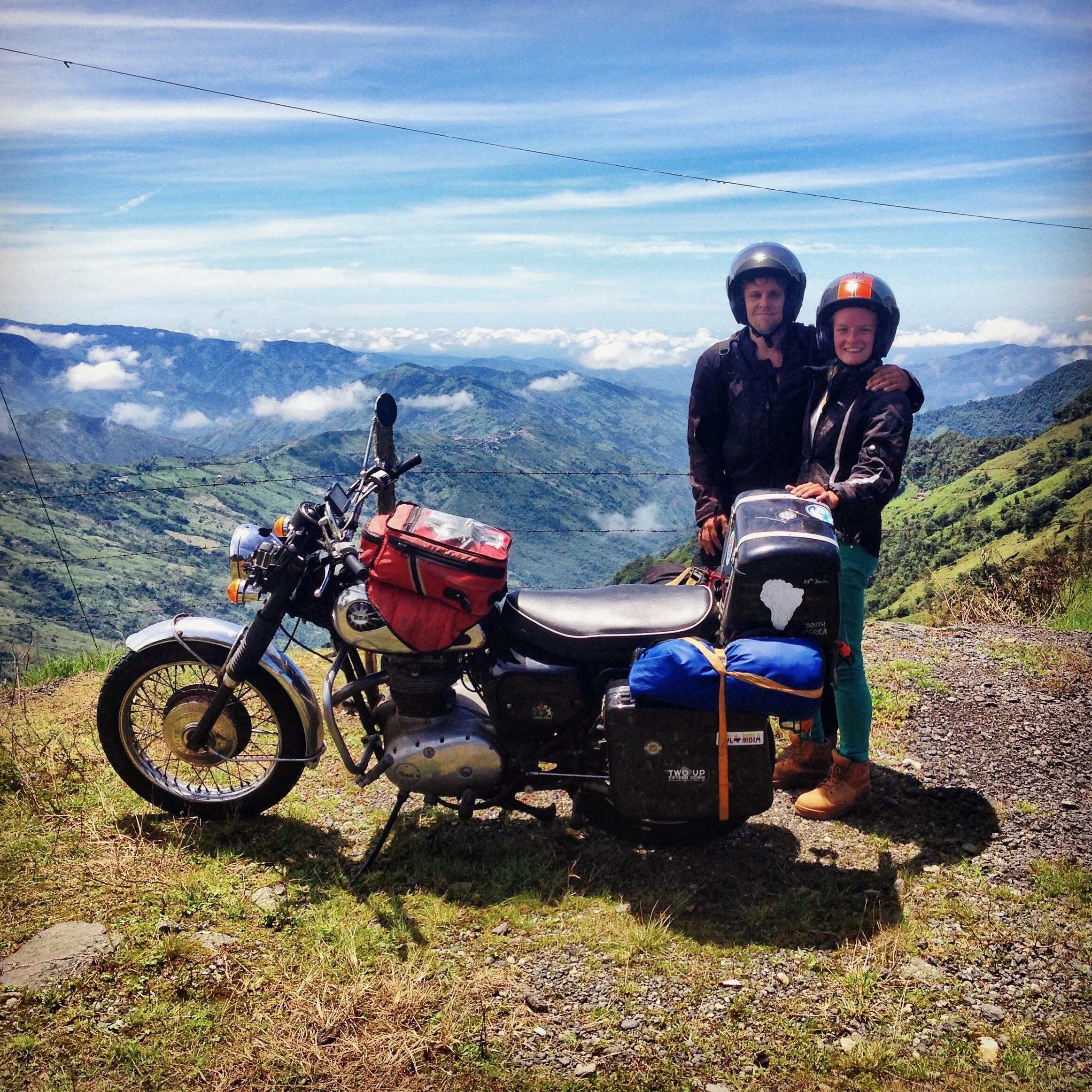 Two people in motorcycle helmets standing outdoors on a mountain with a scenic view of valleys, mountains, and blue sky with clouds, with a motorcycle and luggage in the foreground.