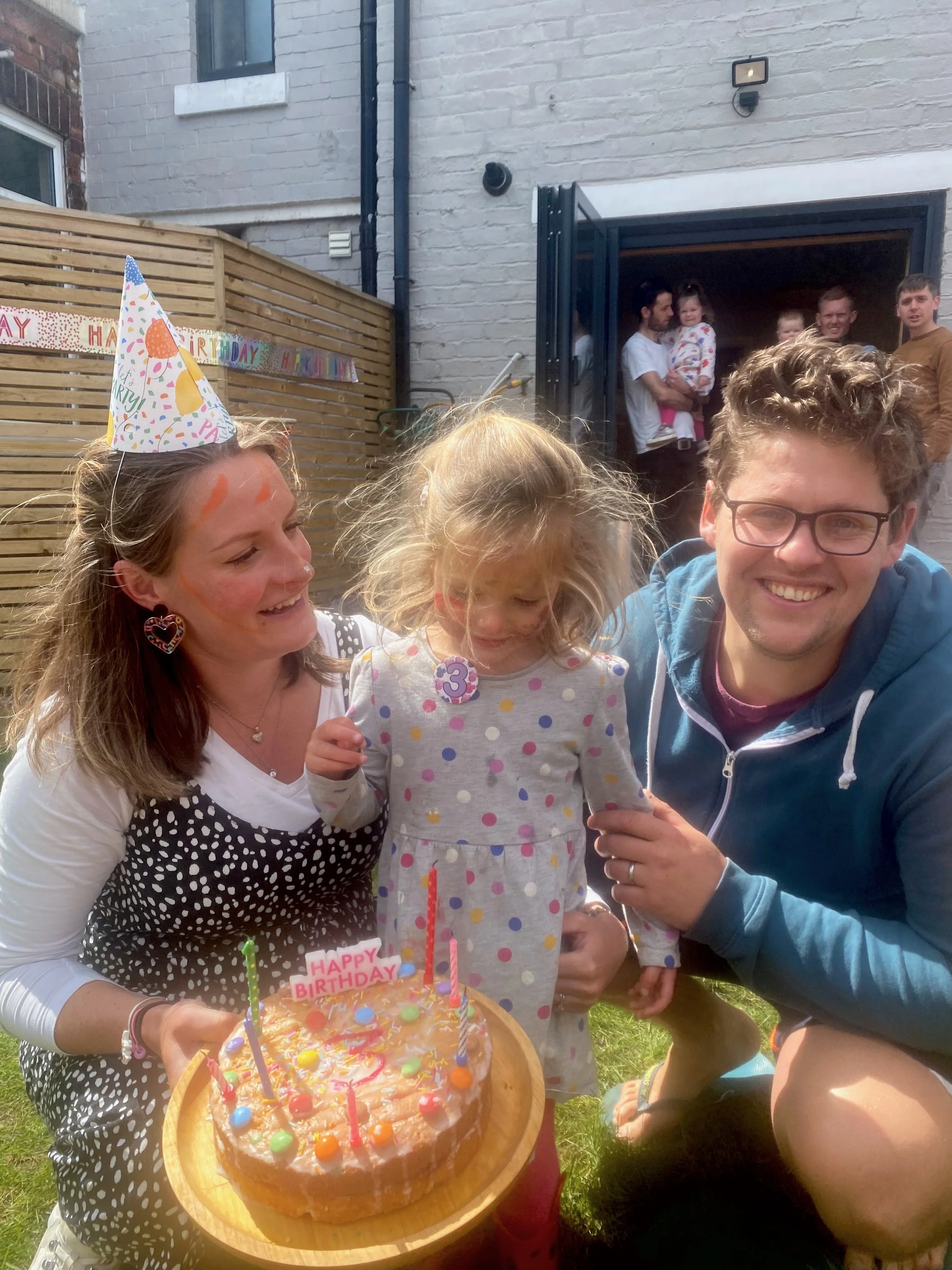 A family celebrating a child's birthday outdoors with a cake that has candles and a birthday sign, surrounded by smiling people, including a woman wearing a birthday hat and a young girl with a '3' badge.