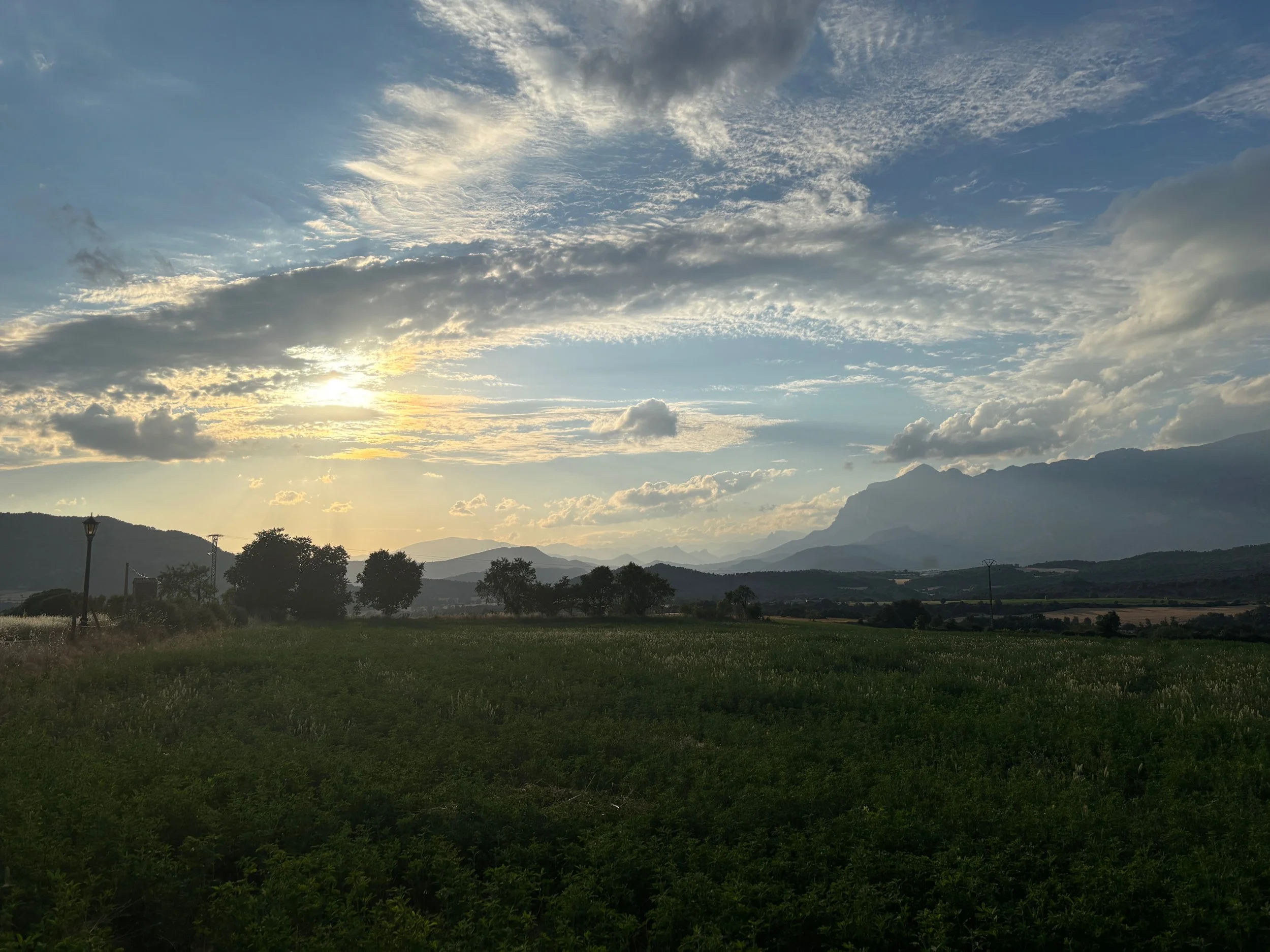A landscape view of mountains in the distance with a partly cloudy sky and the sun setting or rising, casting a glow over the hills and trees in the foreground.