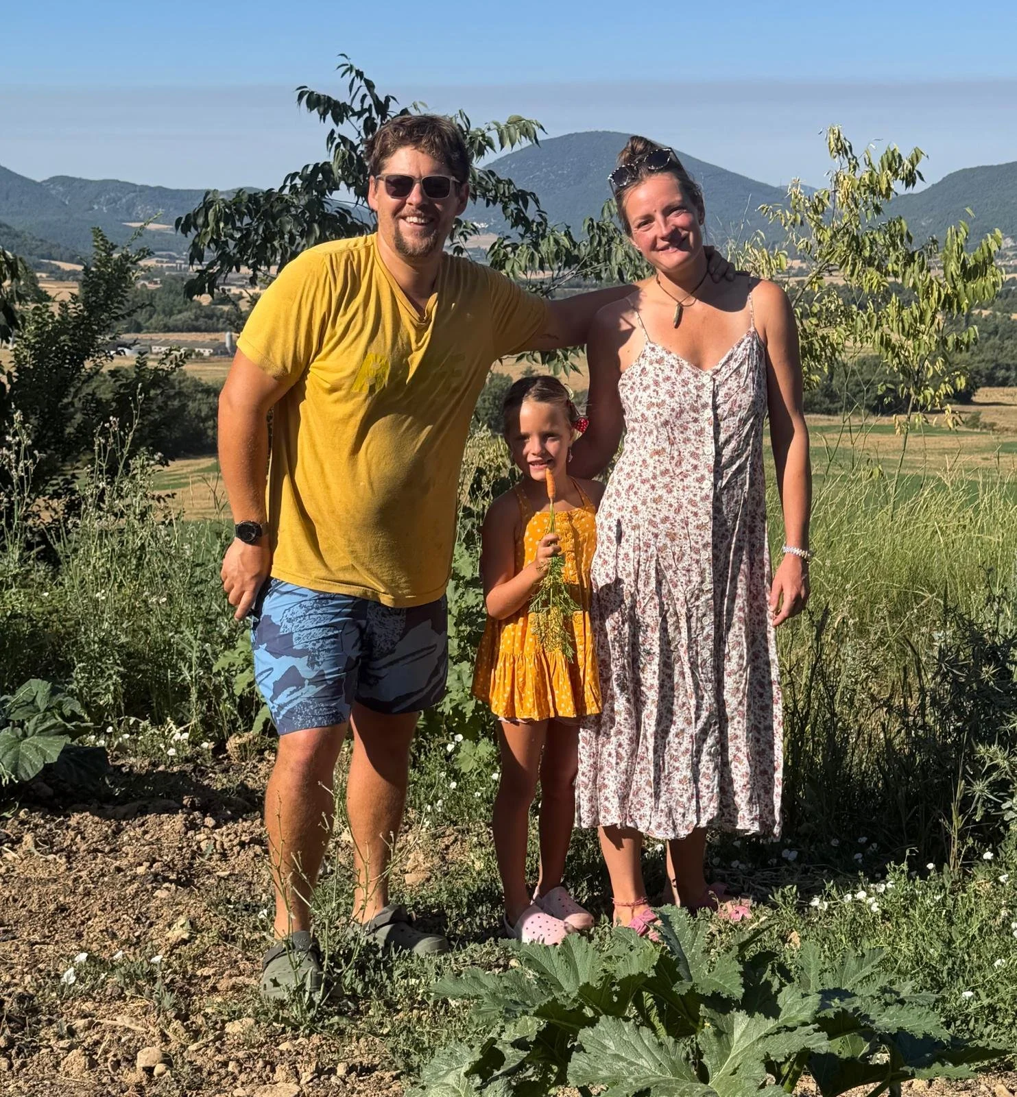 A family of three standing in a garden with mountains in the background, smiling at the camera. The man is wearing sunglasses, a yellow T-shirt, and shorts. The woman is wearing a sleeveless floral dress with sunglasses on her head. The young girl is holding a carrot and wearing a yellow dress with pink shoes.