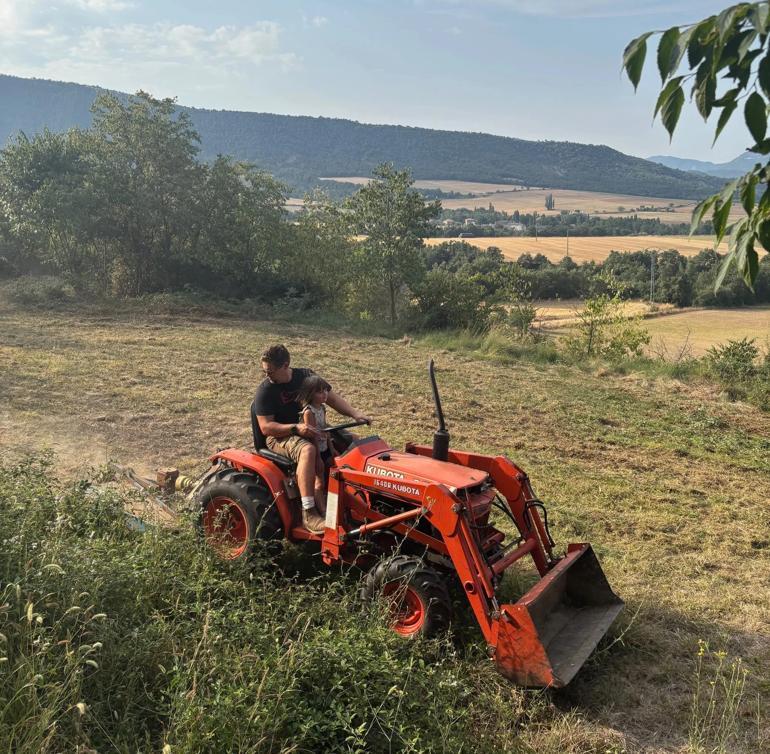 A person and a young girl riding a red Kubota tractor through a grassy field with trees and hills in the background.