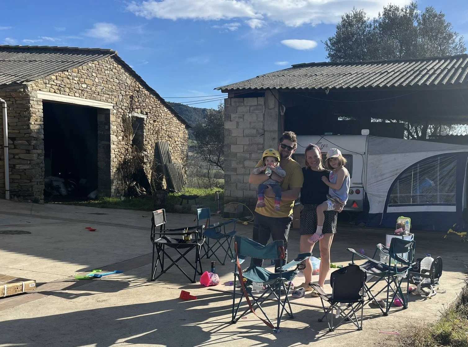 A family of four standing outdoors among scattered chairs and toys. The father, mother, and two young children are smiling, with one child holding a water bottle and wearing a yellow helmet. The setting appears to be a backyard or a rural area with stone and brick structures and a blue sky.