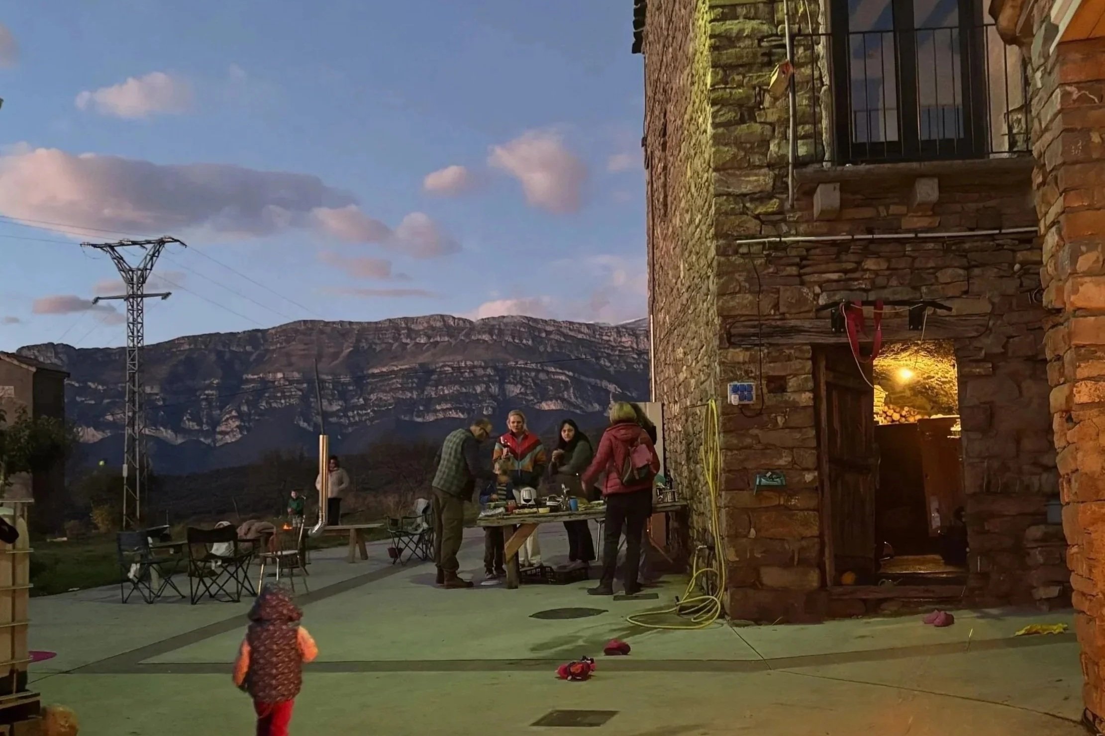 A group of people gathered outdoors in the evening, with a mountain range and a blue sky with clouds in the background. The scene includes a brick building with a small balcony, a table with items on it, and a woman and children playing on a green surface. There are chairs and some objects scattered around, and an electric power pole is visible in the distance.