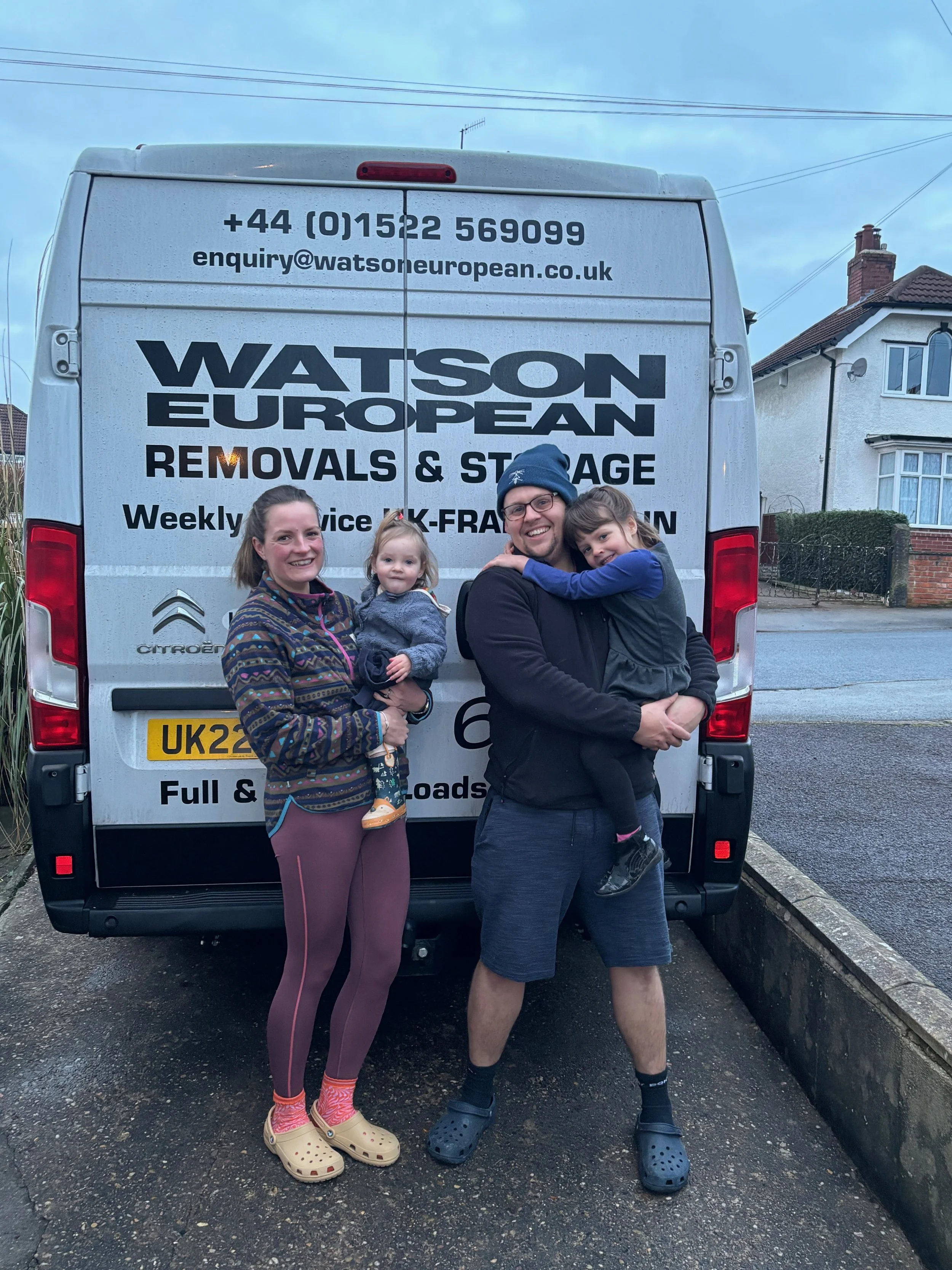 A family of four standing in front of a Watson European removals van, smiling. The family includes a woman, a man, and two young children. The woman is holding a toddler, and the man is holding an older child. The van has contact information and company details displayed on the back.