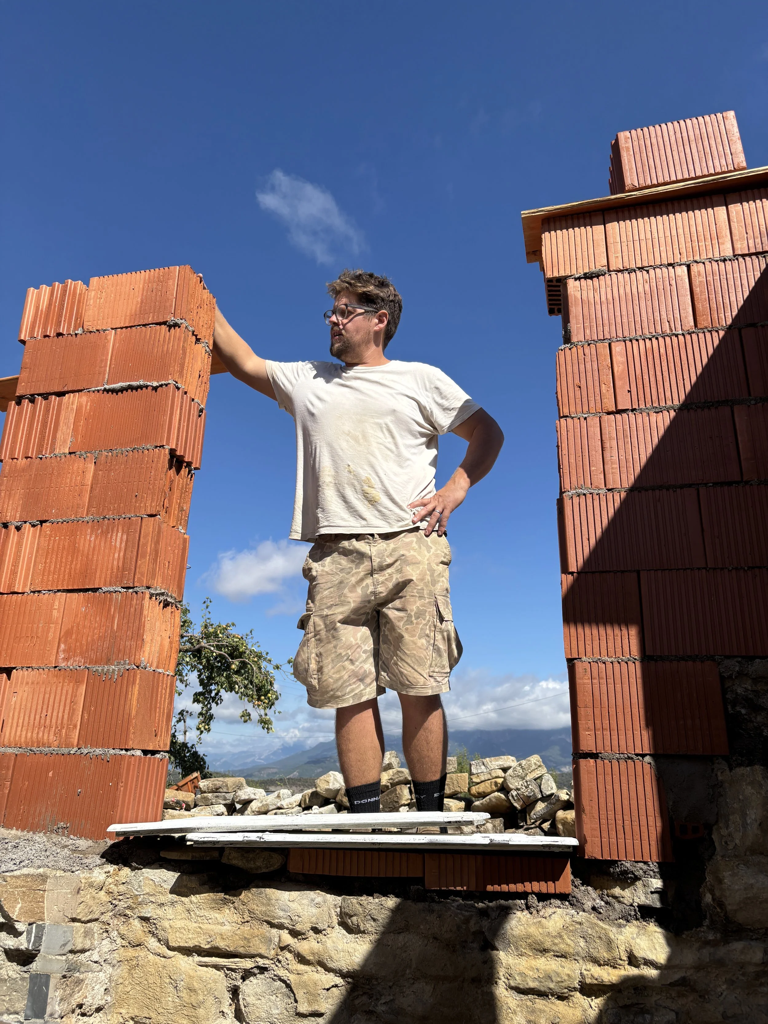 A man with glasses and a beard wearing a white t-shirt and camouflage shorts standing on a plank in front of brick walls, with mountains and blue sky in the background.