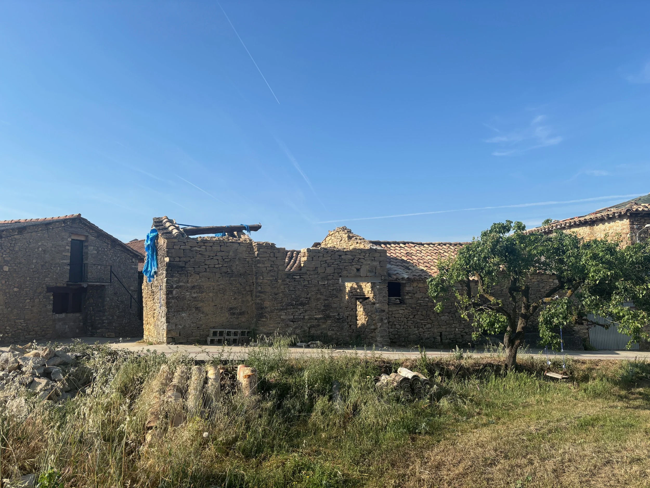 Old stone buildings with tiled roofs, one building partially collapsed, a blue cloth hanging from a wooden beam, a tree on the right side, and a grassy area with rocks and debris in the foreground under a clear blue sky.