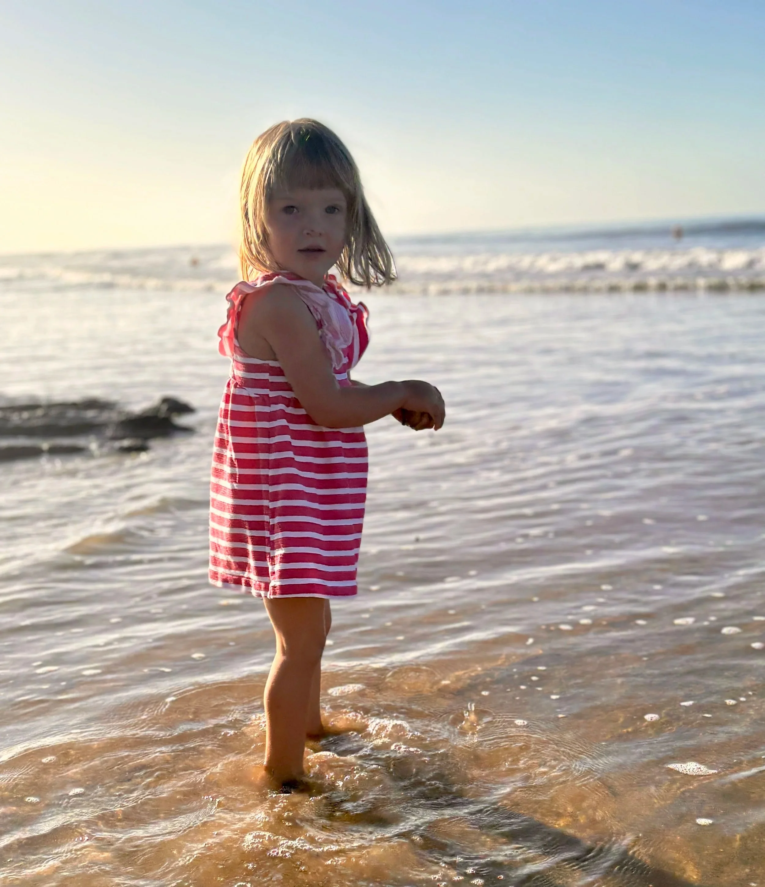 Young girl with shoulder-length hair wearing a pink and white striped dress standing in shallow ocean water at the beach, facing the camera with a surprised expression, during sunset.