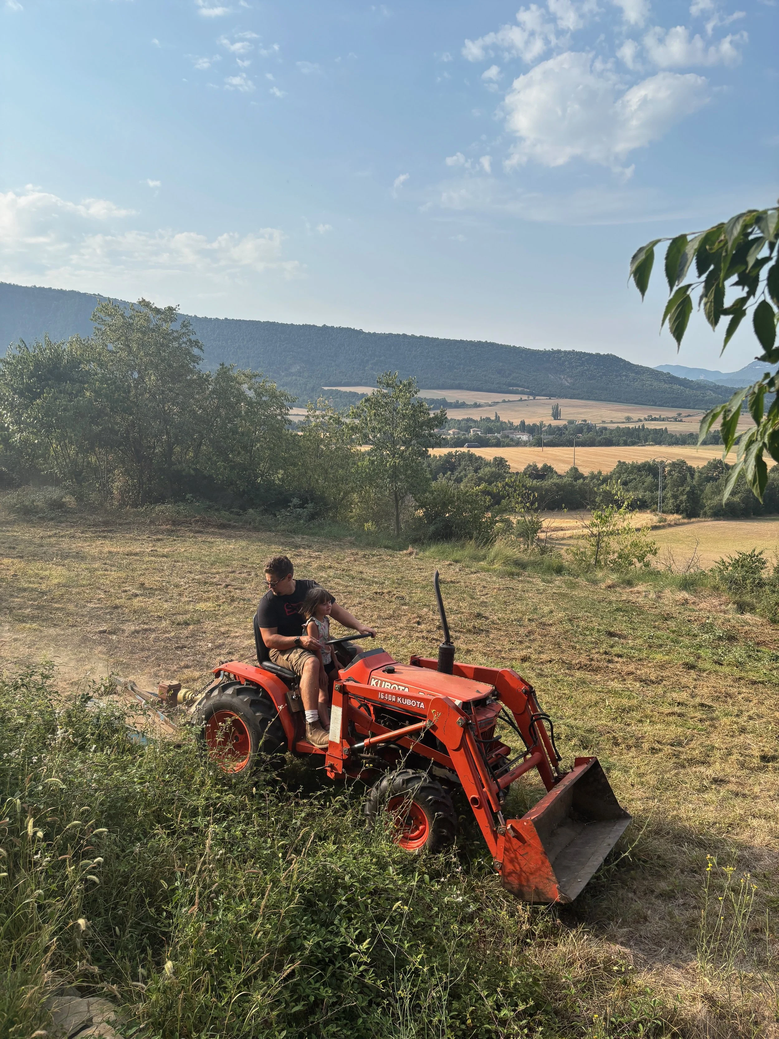 A man and a young girl riding a small orange tractor through a grassy field, with hills and a blue sky with clouds in the background.