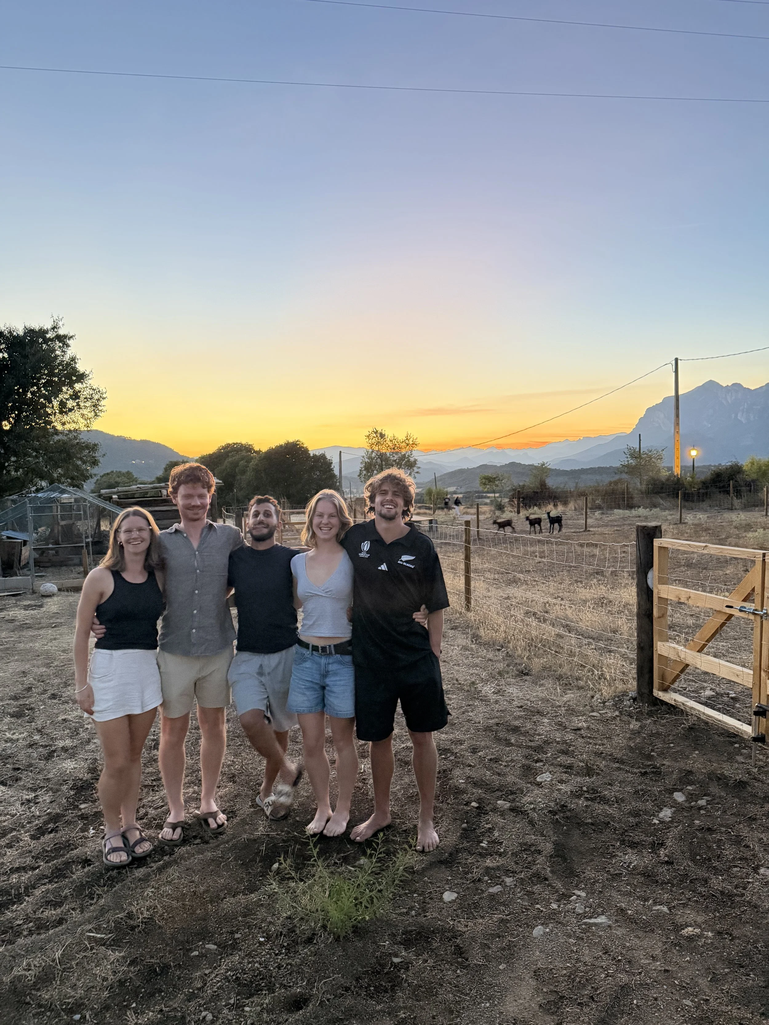 Group of six young adults standing outdoors during sunset, smiling, with mountains in the background and a cloudy sky.