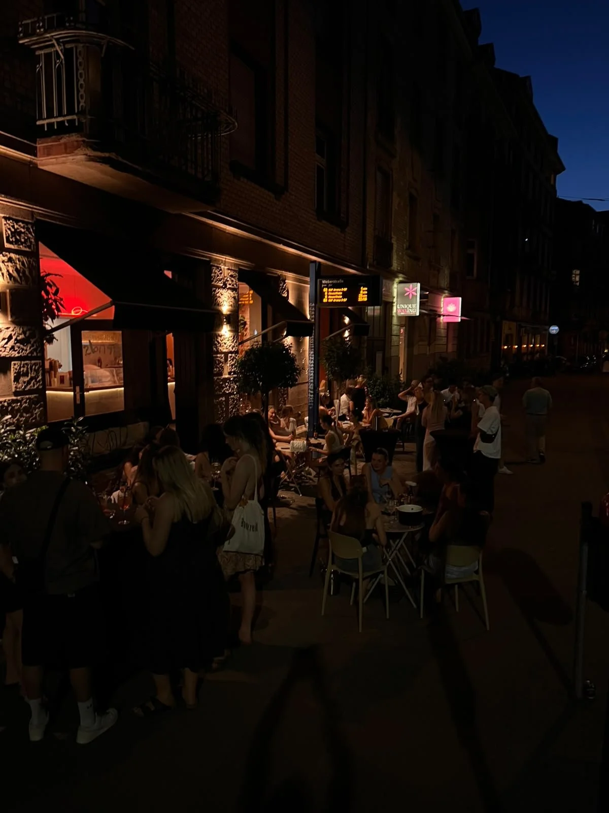 Menschen sitzen abends draußen vor einer Bar oder einem Restaurant, das mit warmem Licht beleuchtet ist, an Tischen und genießen die Gesellschaft, während das Stadtbild im Hintergrund zu sehen ist.
