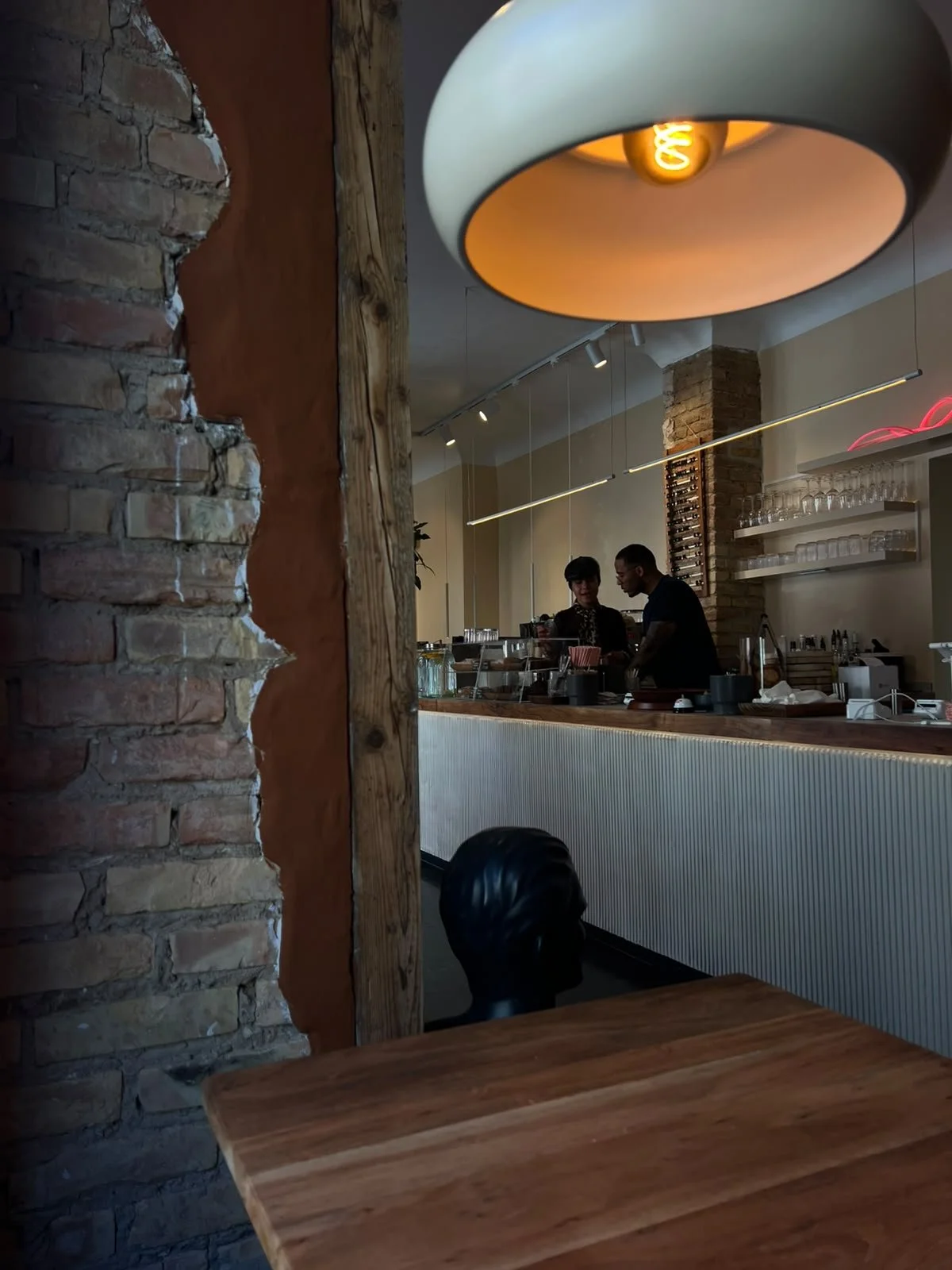 Blick auf eine Bar mit zwei Menschen hinter dem Tresen, einer Frau und einem Mann, in einer modern gestalteten Einrichtung mit alten Ziegelwänden, Holzregalen und warmem Licht. Im Vordergrund ist ein leerer Tisch zu sehen.