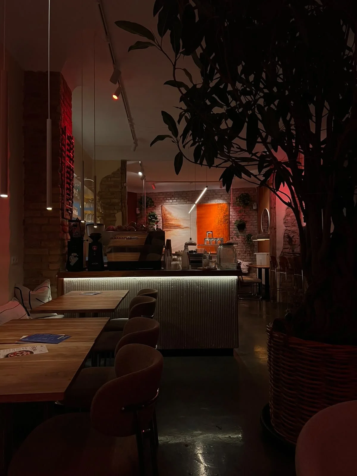 Innenaufnahme eines gemütlichen Cafés mit Holz- und Backstein-Elementen, weichem Licht und stilvoller Dekoration, inklusive Tischen, Stühlen, einer Theke und Pflanzen.