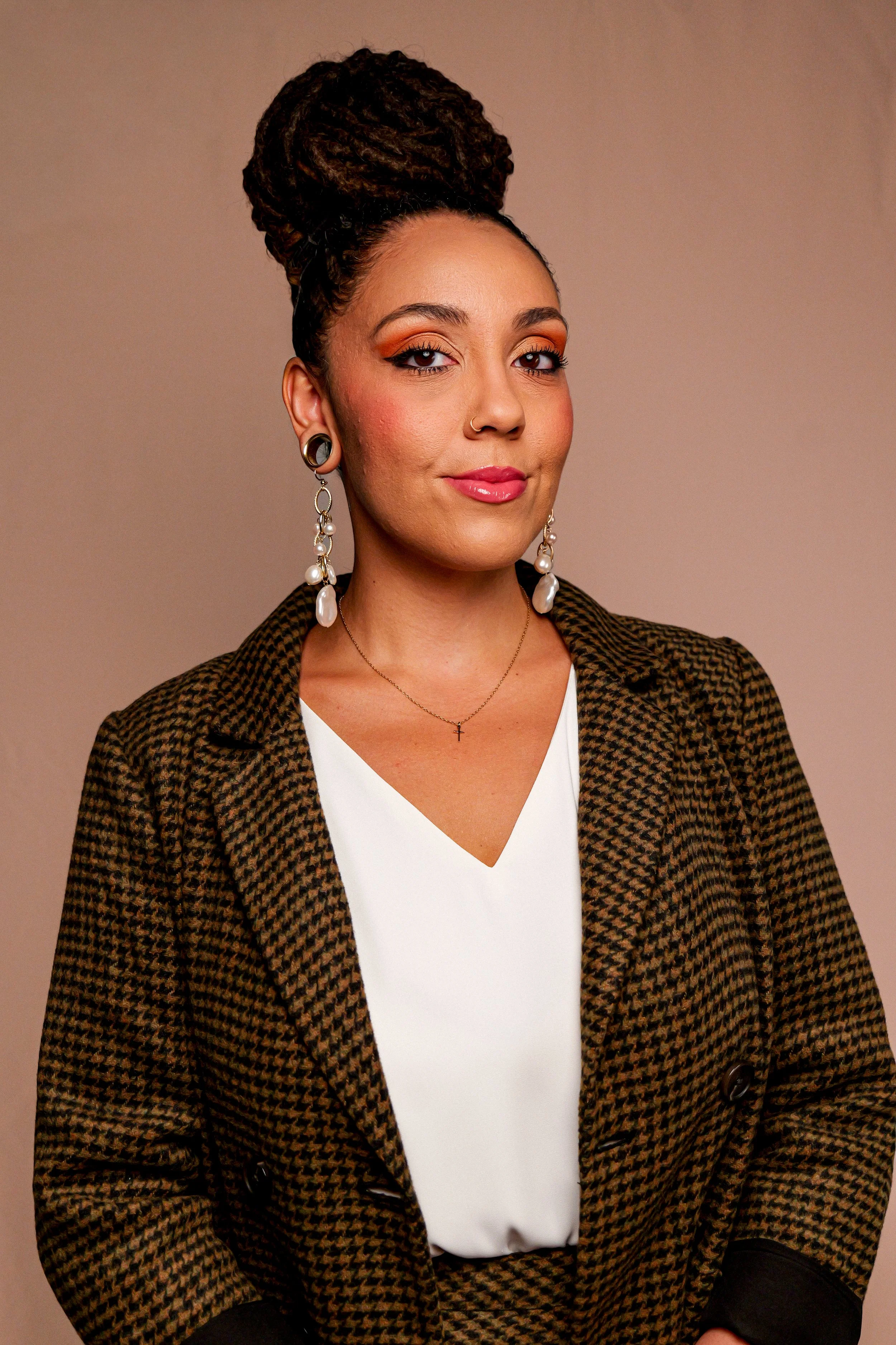 A woman with medium skin tone and dark brown hair styled in a large braided bun. She has makeup with orange eyeshadow and pink lipstick, wearing large pearl earrings, a delicate necklace with a small cross, a white V-neck top, and a houndstooth blazer. She is posing with a neutral background.