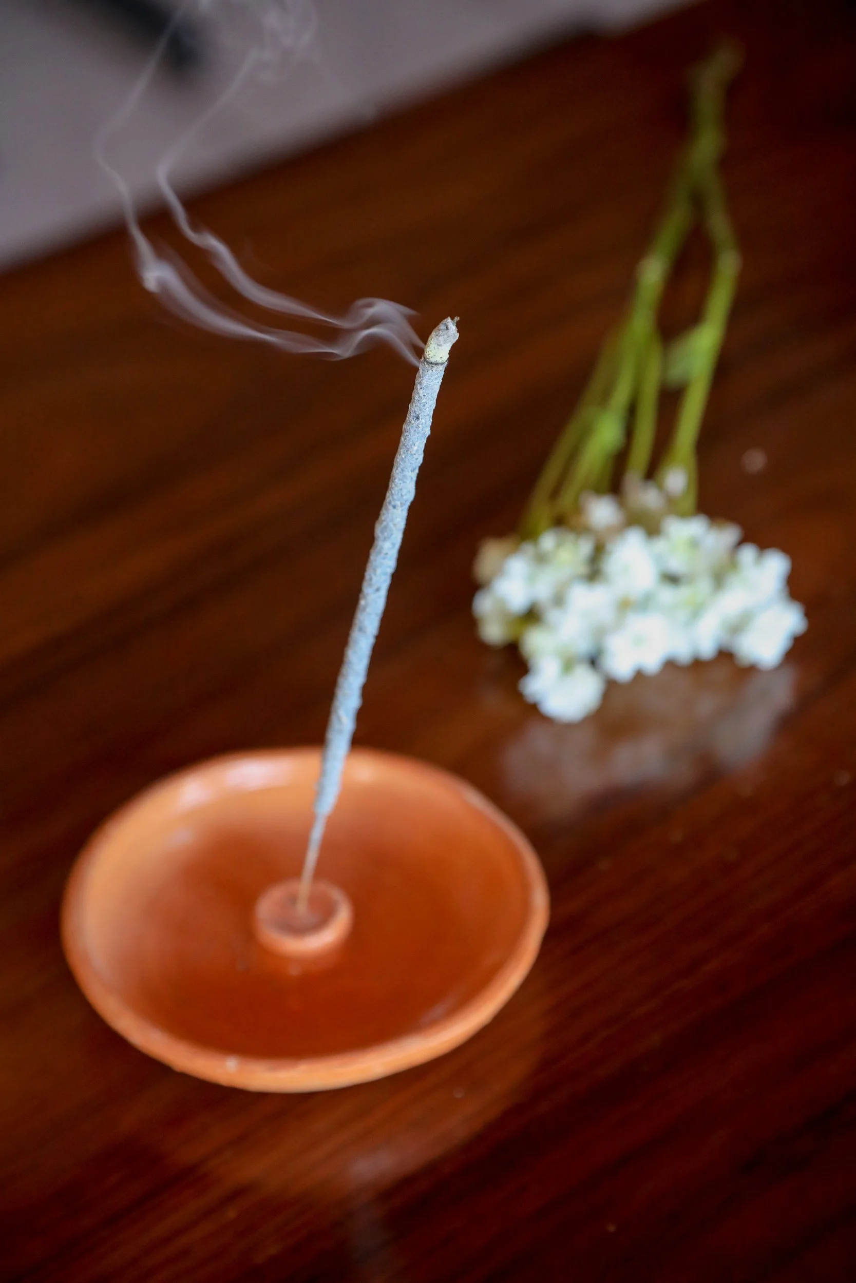 A lit incense stick with smoke rising from its tip, placed in a small terracotta holder, with a blurry flower in the background on a wooden surface.