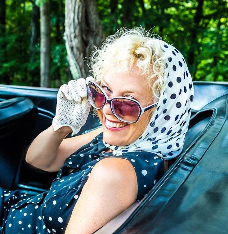 A smiling woman with curly blonde hair, wearing sunglasses, a polka dot dress, a polka dot headscarf, and a white dotted glove, sitting in a vintage convertible car outdoors with green trees in the background.