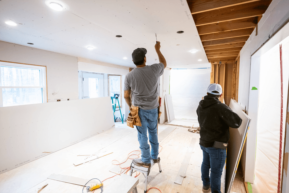 Two construction workers installing a ceiling panel inside a house under renovation. One is standing on a step stool, reaching up with a tool, while the other holds a panel.