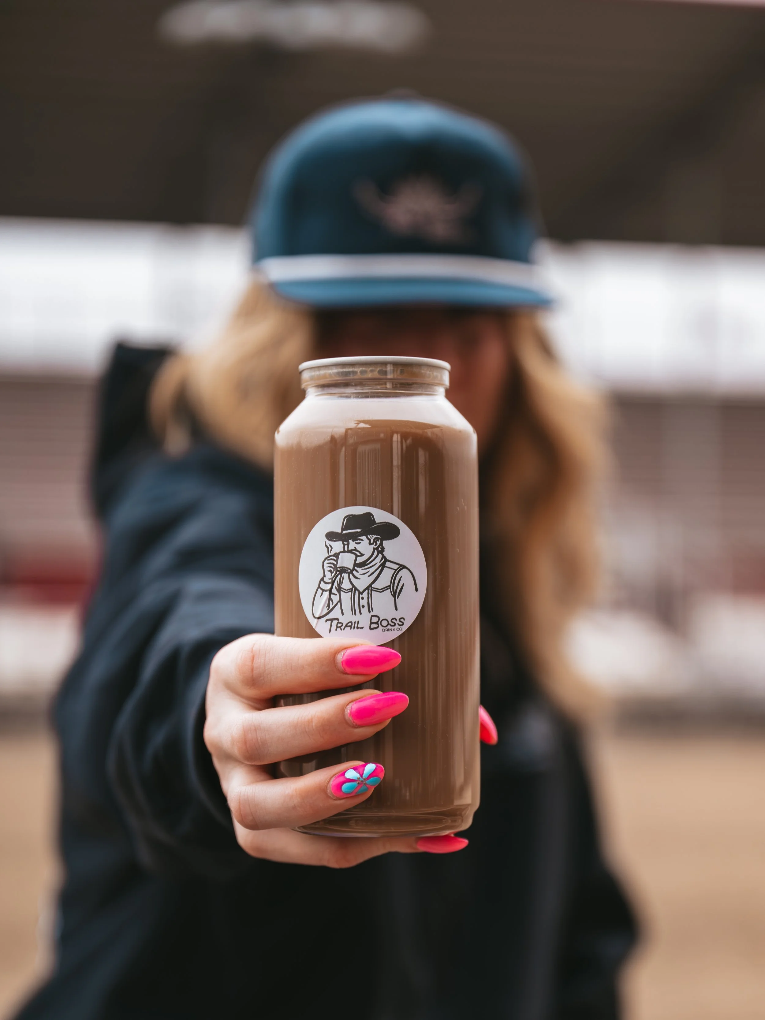 Person holding a bottle of chocolate milk with a 'Trail Boss' logo, showing a man wearing a hat and drinking from a cup, with purple nails that have a colorful flower design.