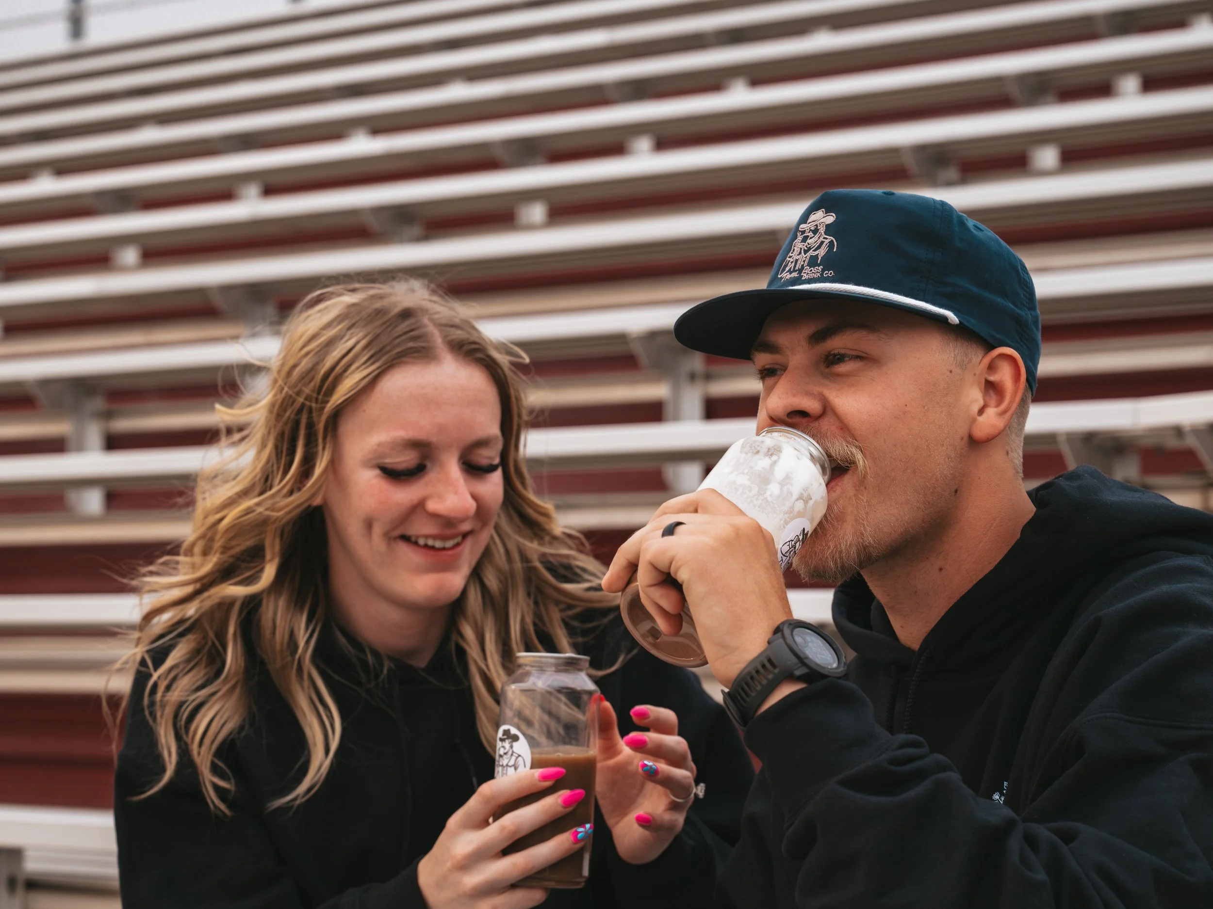 A young woman and a young man enjoying drinks together outdoors, with stadium seating in the background.