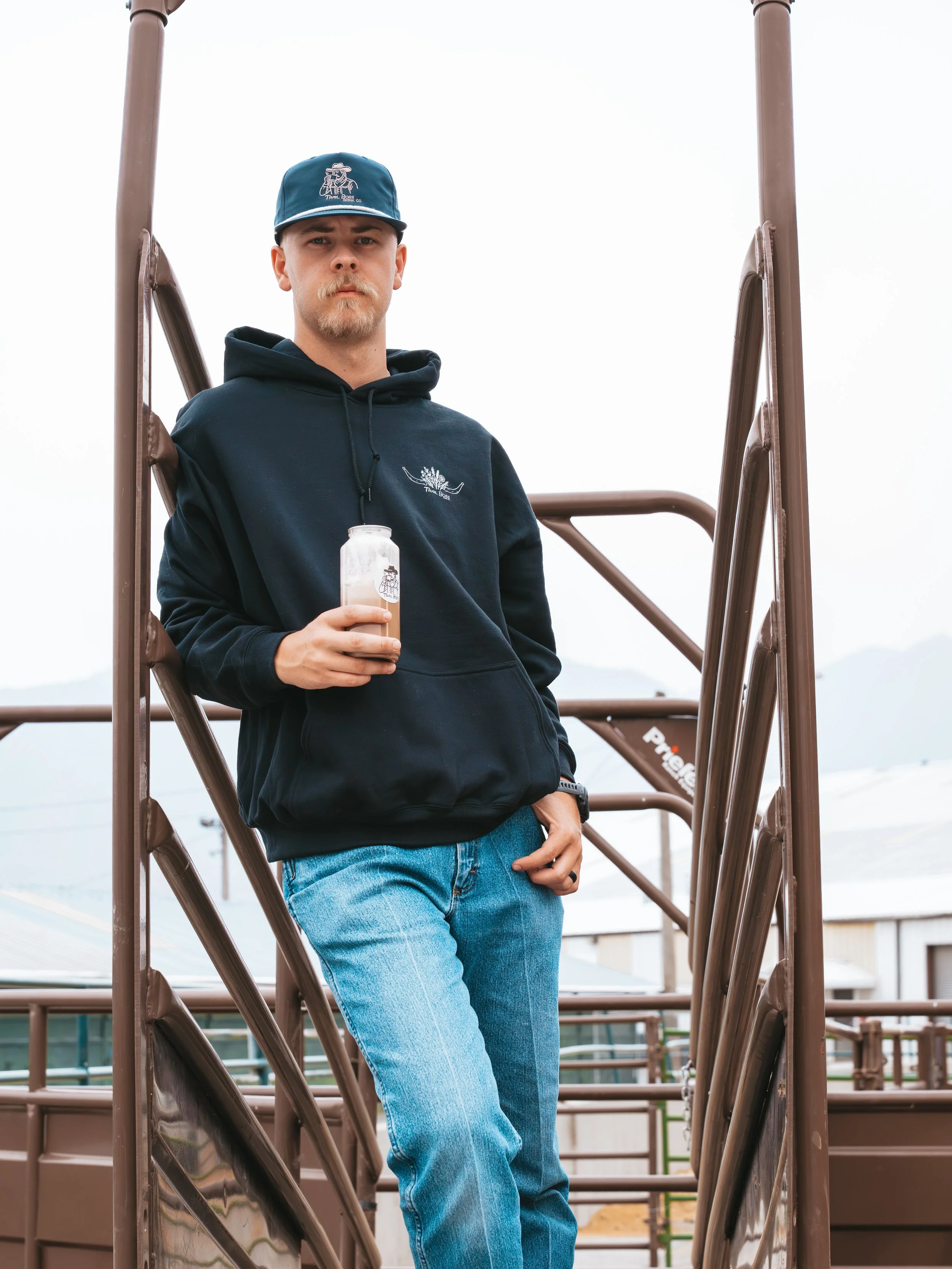 A young man with light skin, a goatee, wearing a black hoodie with a graphic print on the front, blue jeans, and a blue baseball cap, stands on a playground slide holding a drink, with an overcast sky in the background.